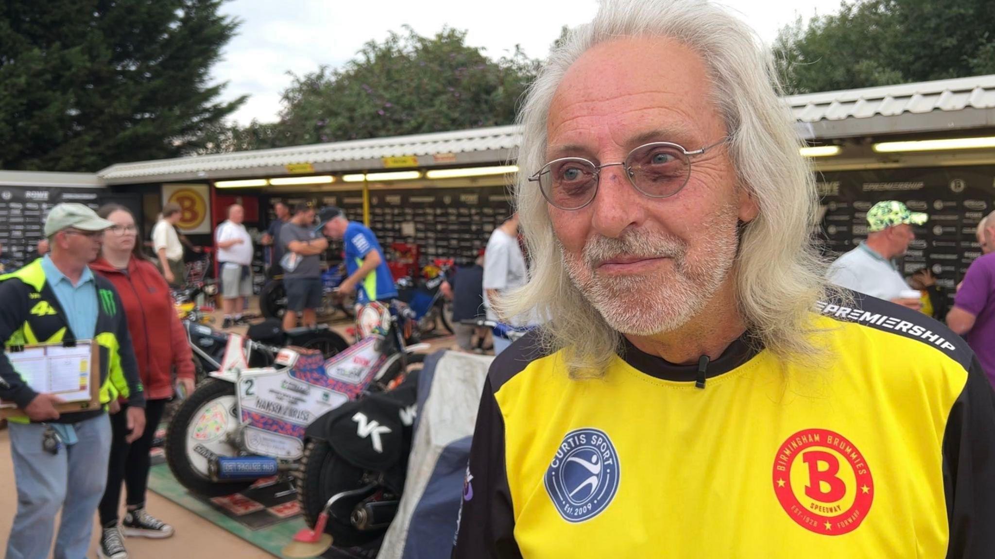 Nigel has long white hair and circular silver glasses. He is wearing a black and yellow t-shirt with a large red logo with the letter B on it. Behind him are speedway bikes parked up in rows, with people looking at them