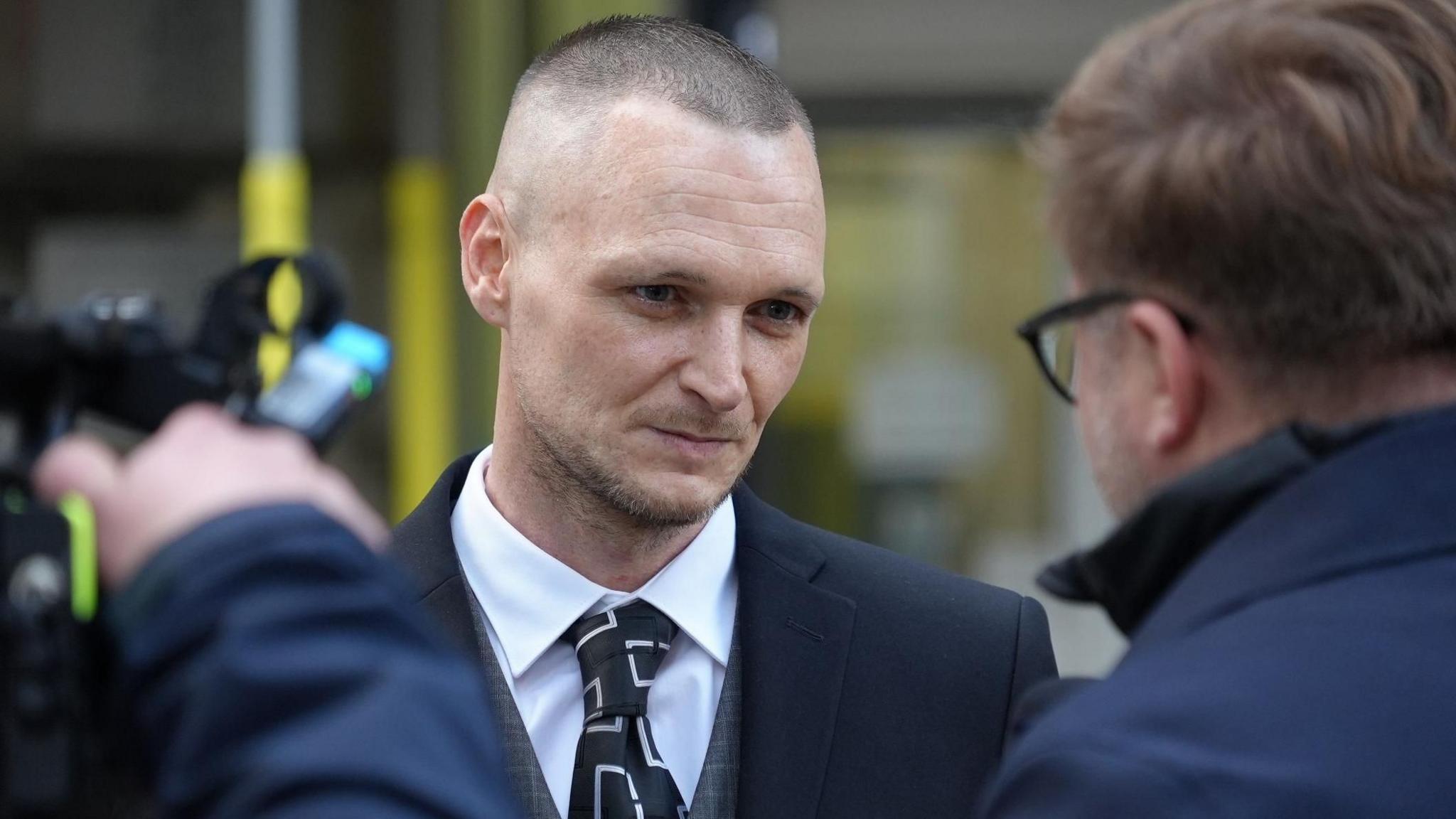 James Howells listening to reporter who is facing him, with a camera man to the left of the reporter. Mr Howells is wearing a white shirt and black tie with a white pattern and a grey checked waistcoat and navy blazer.