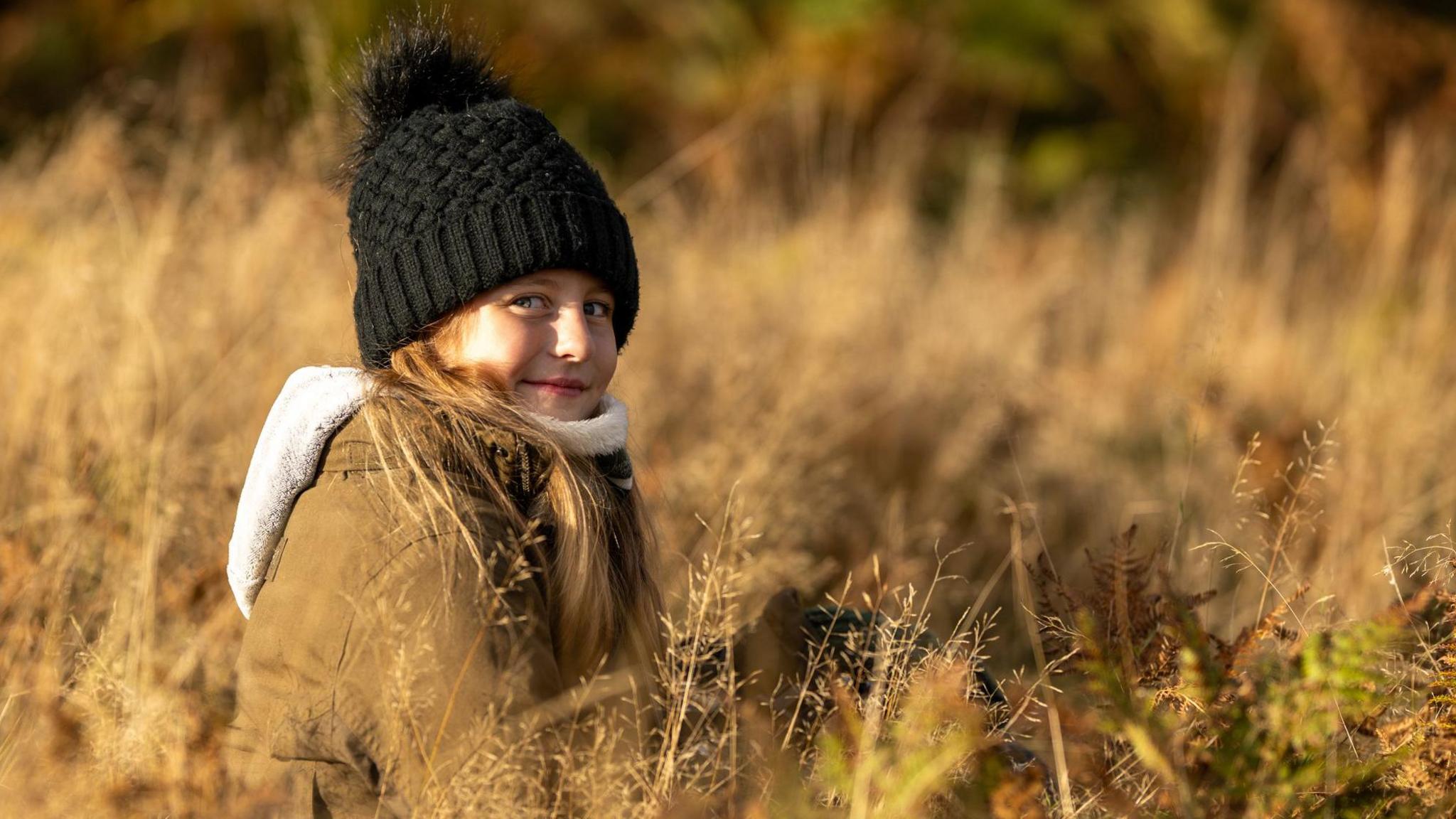 Photograph of 10-year-old Jamie, among the yellow grass. She wears a parka jacket with a fur trim, and wears a black bobble hat. She has long brown hair and blue eyes. There is an orange haze over the image.