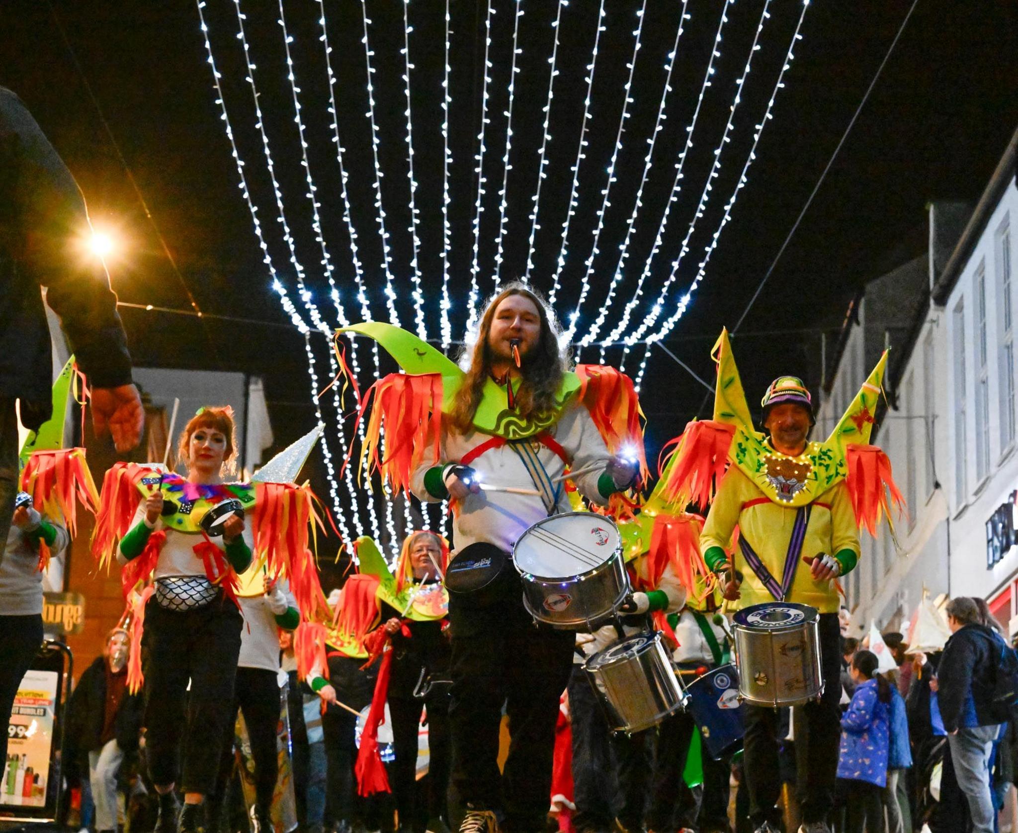 Drummers in bright yellow and orange costumes walk along under a string of lights