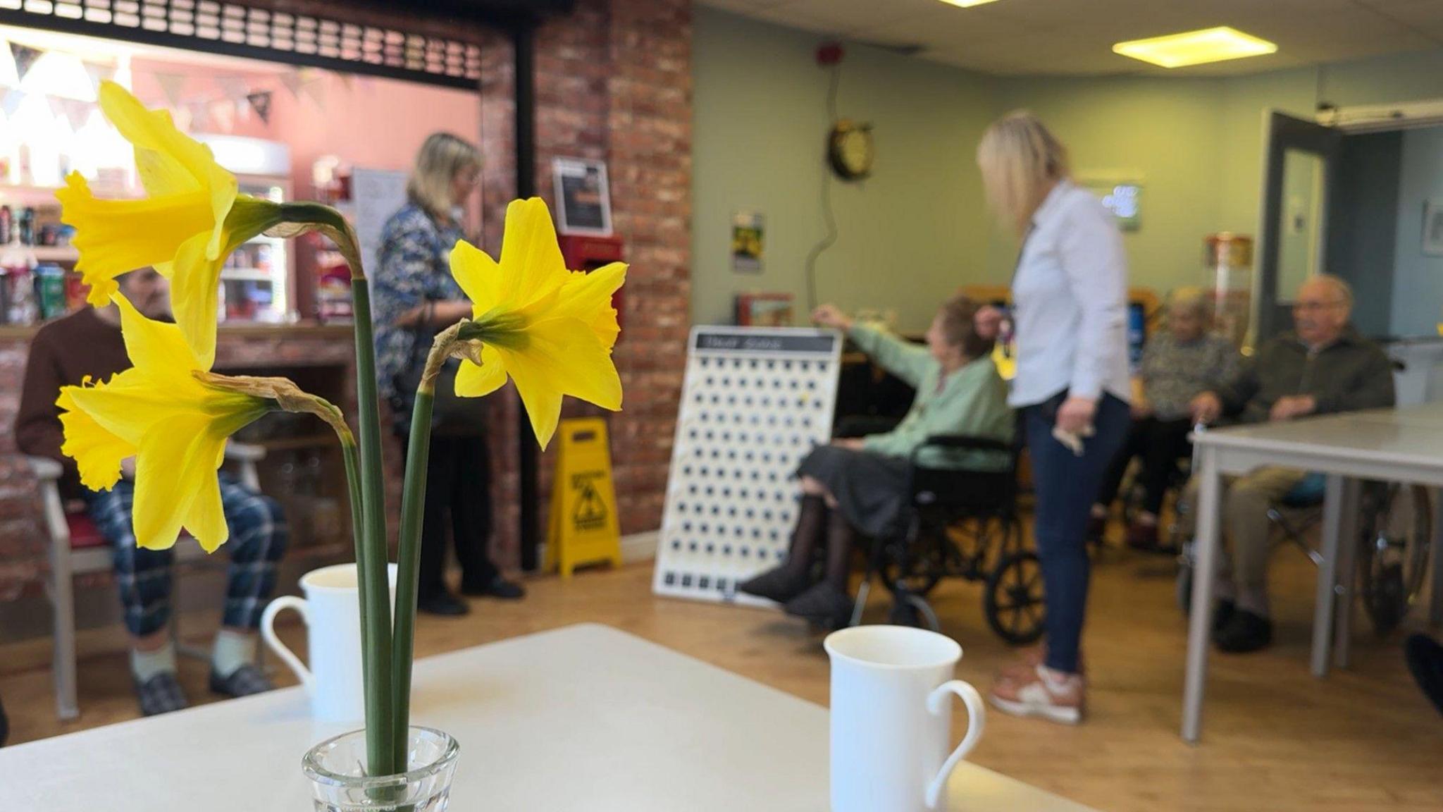 Daffodils are in the foreground of the image, with care home residents playing games and drinking coffee. 