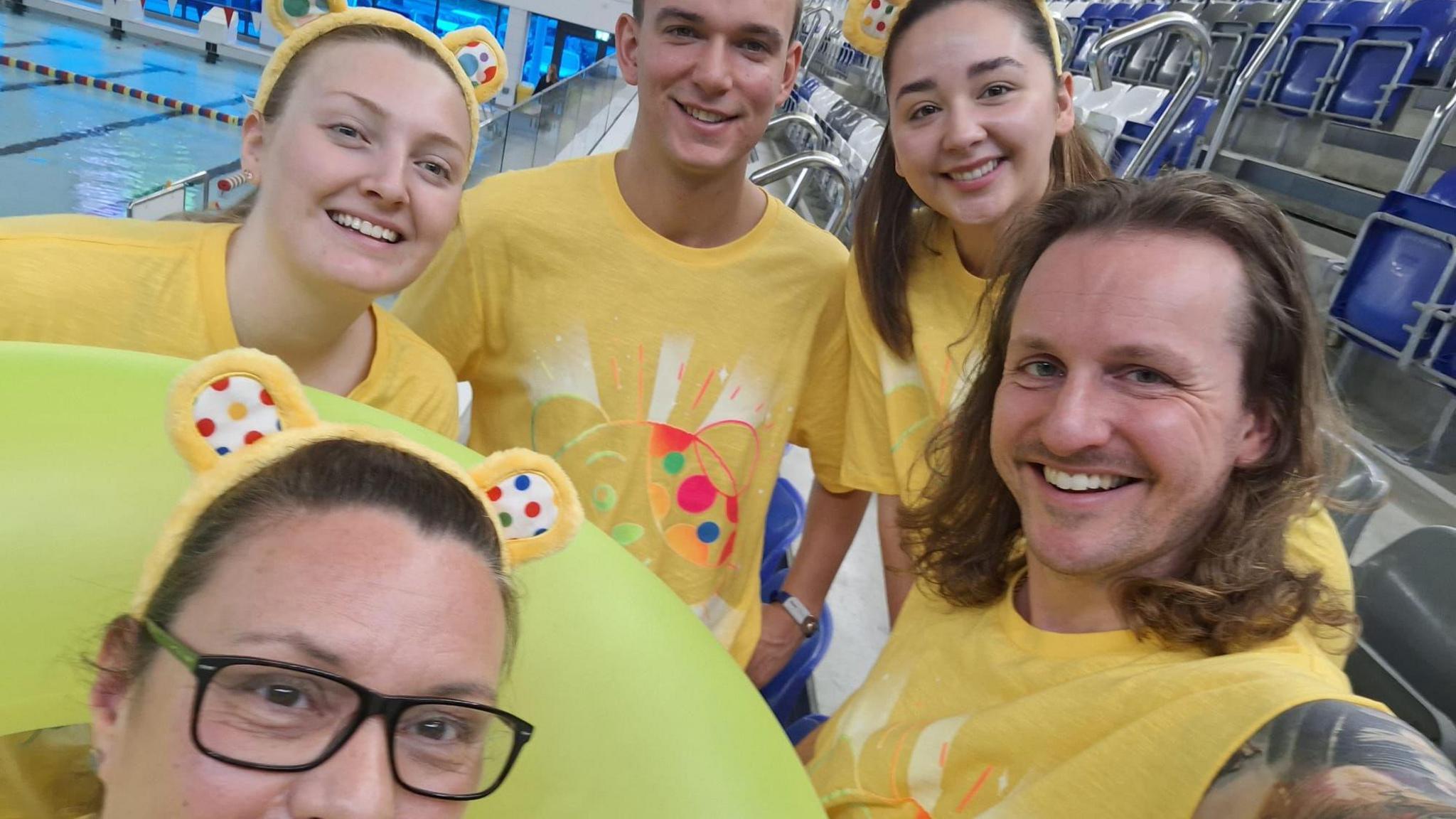 A group selfie photo of the Essex swimming team, they are smiling and wearing yellow shirts, three of them are wearing yellow and polka dot Pudsey ears