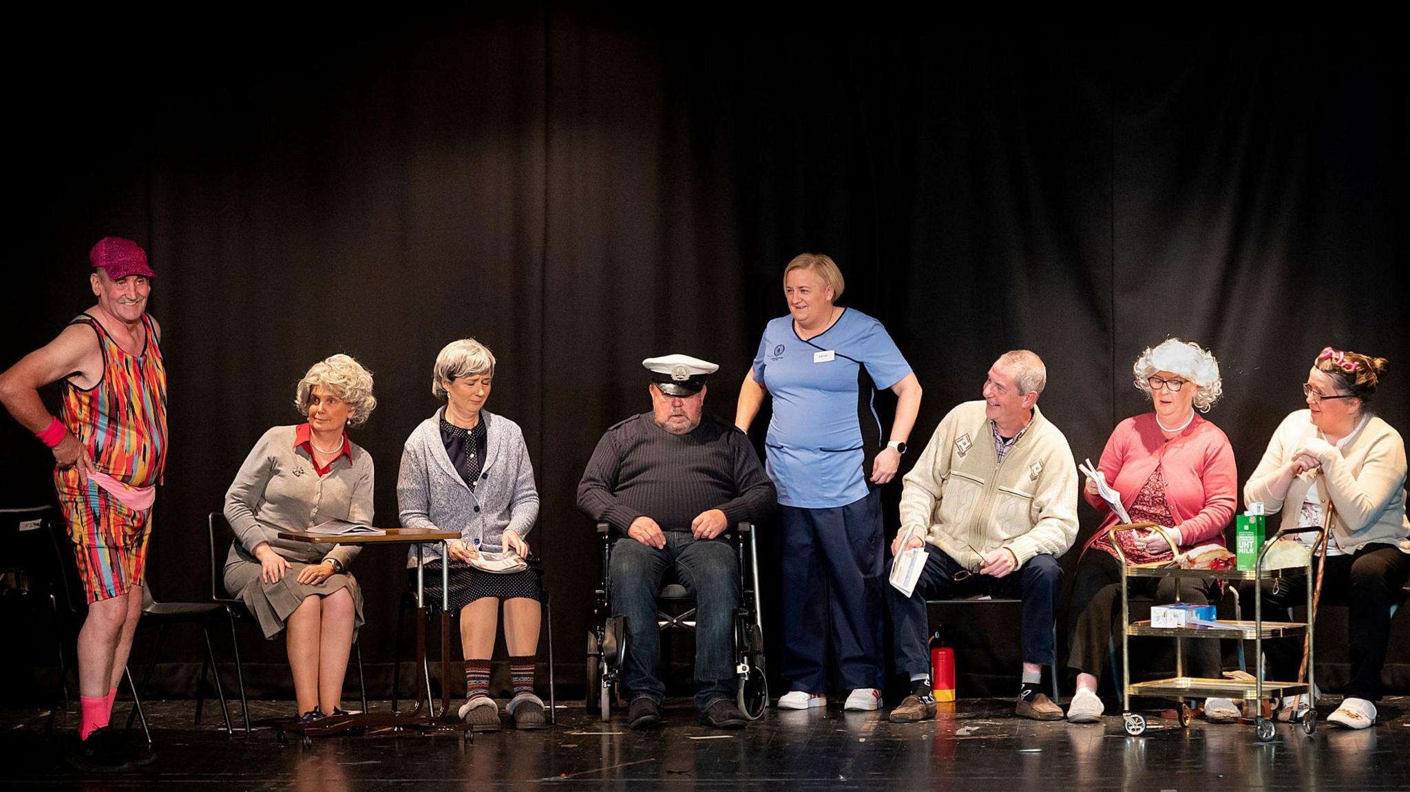 The drama group are on stage wearing an assortment of costumes including sportswear, seaman's cap and nurses uniform.