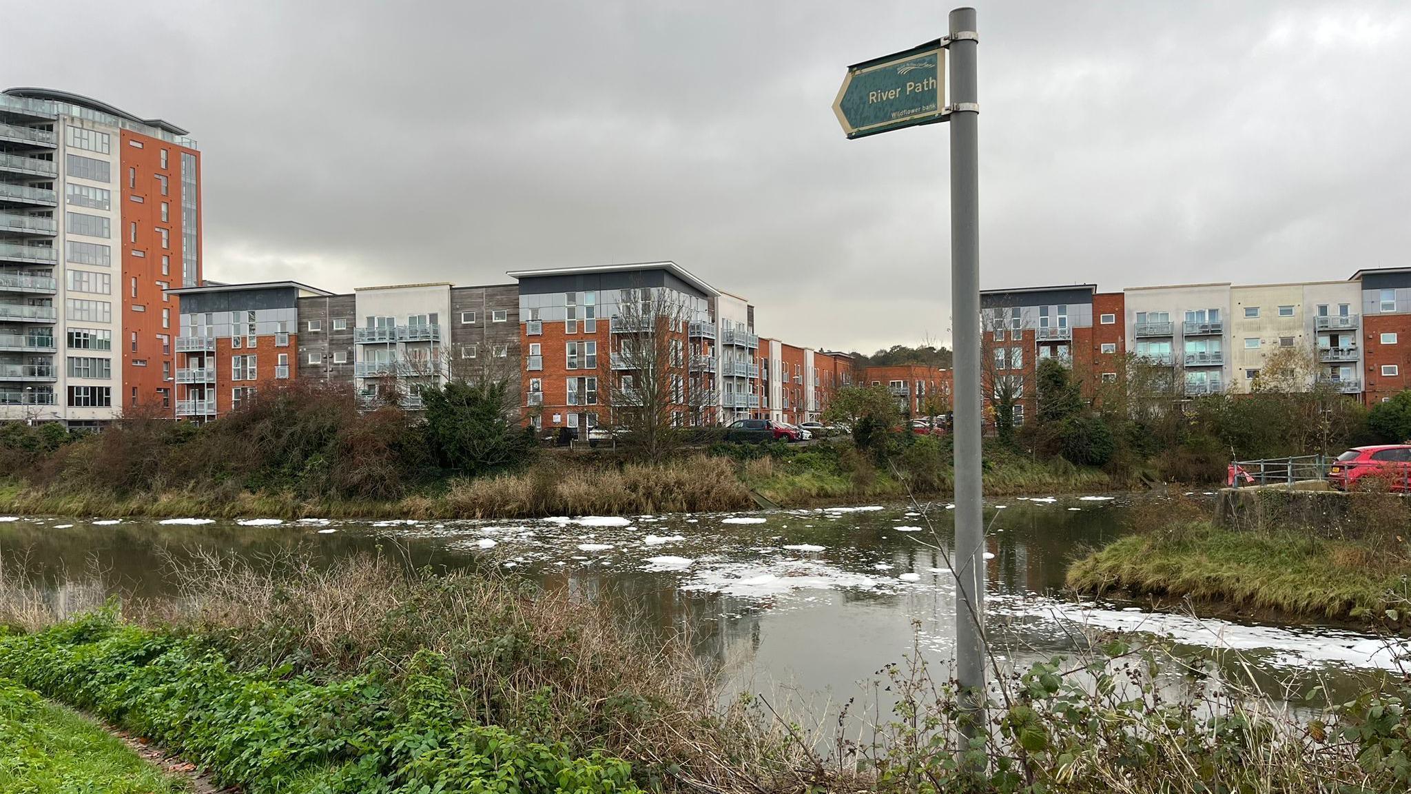 A river with spots of white foam on it during a grey and cloudy day. Blocks of flats can be seen in the distance. A sign detailing the footpath next to the river stands in front of the river. 