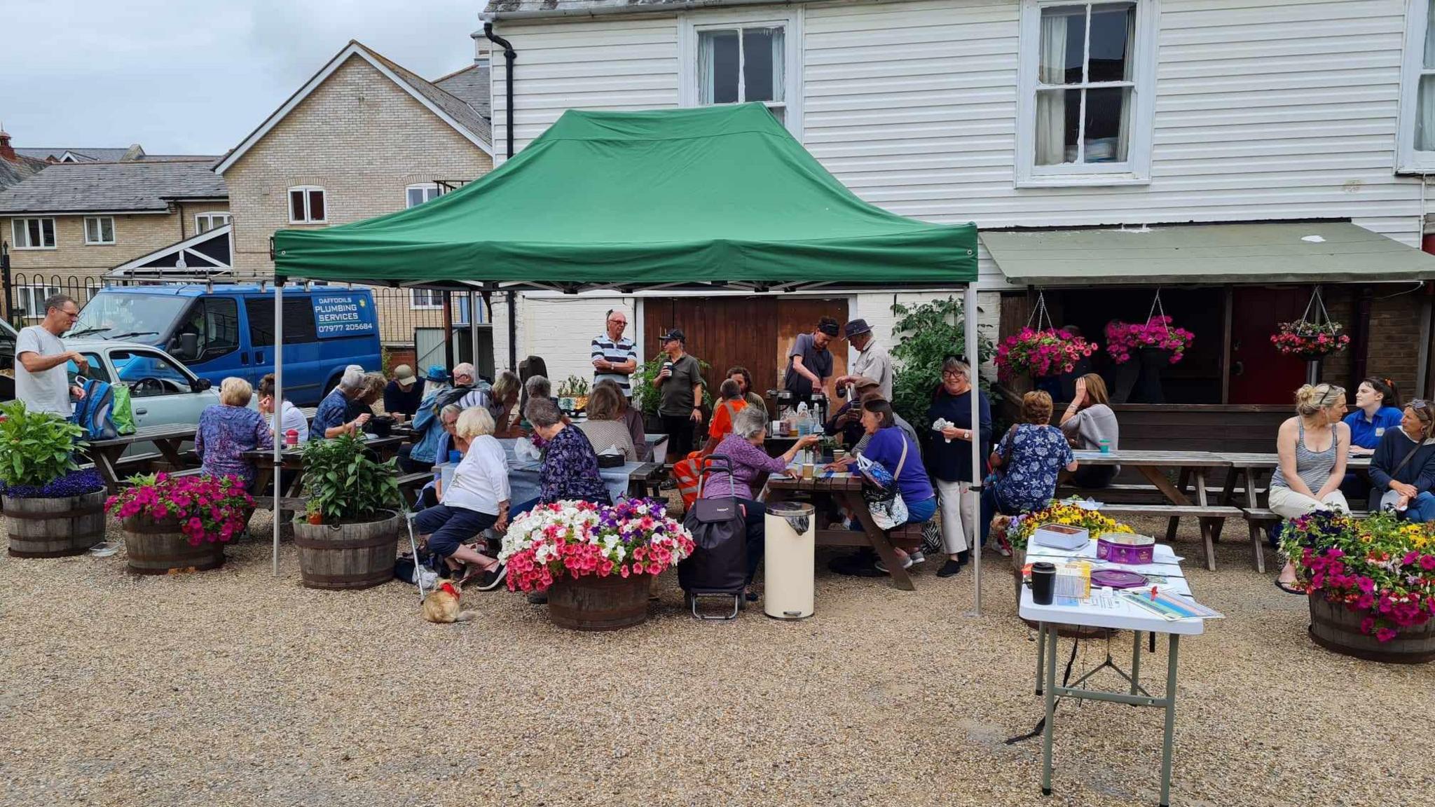 A crowd of people sat on benches outside a pub with lots of planters full of flowers