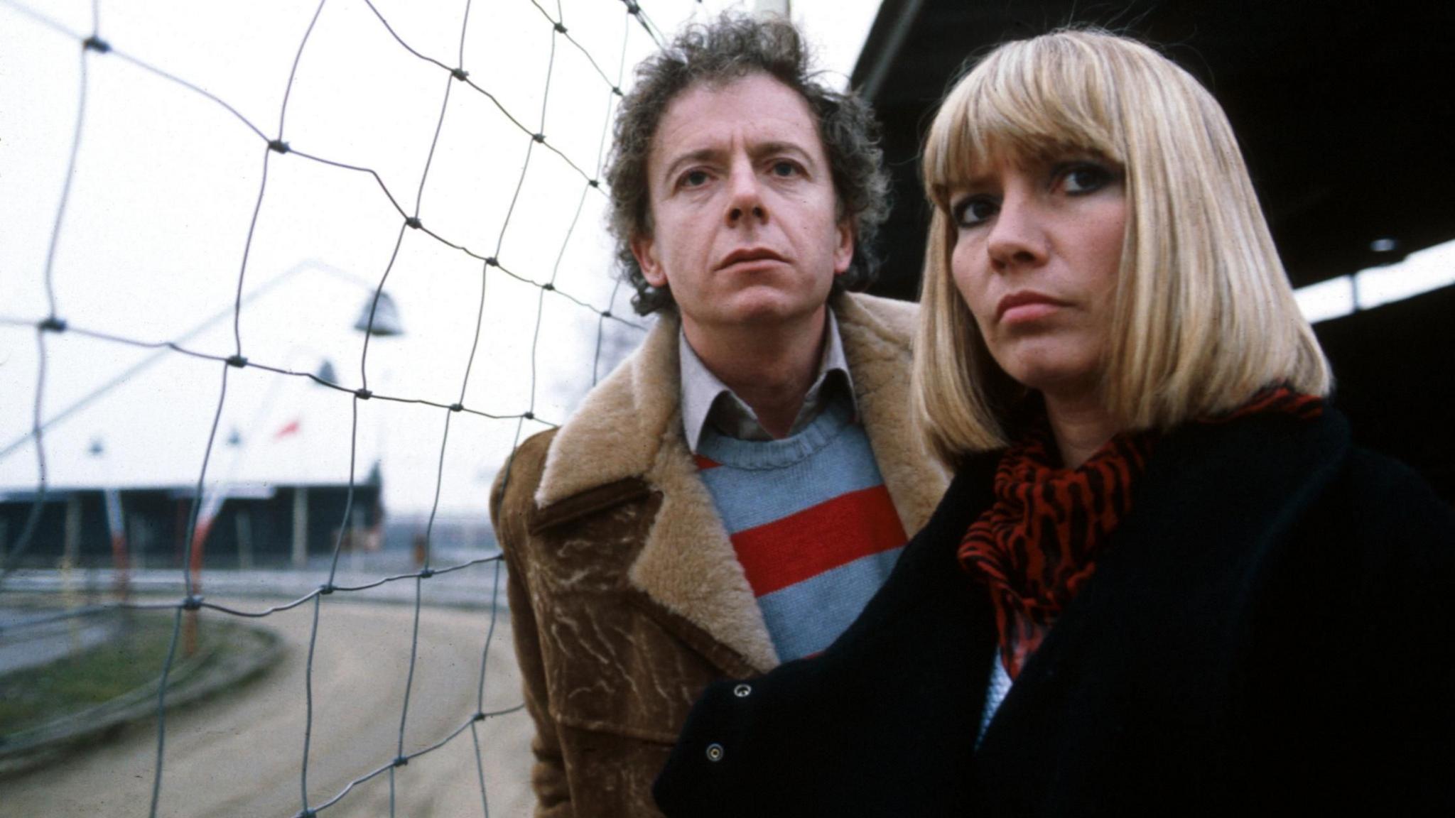 Ray Brooks and Sharon Duce in character on the BBC programme Big Deal. Brooks wears a brown shearling jacket above a striped blue and red jumper. Duce has a short blonde bob and wears a large black jacket with a red and black patterned scarf.