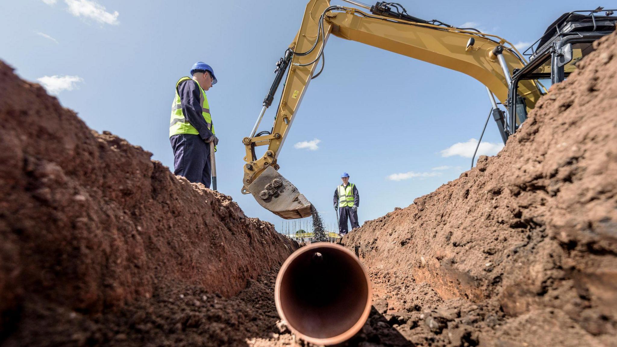 work men in high-vis vests and blue hard hats are stood around a ditch. In the ditch is a brown water pipe which is being buried by earth dropped by a yellow digger.