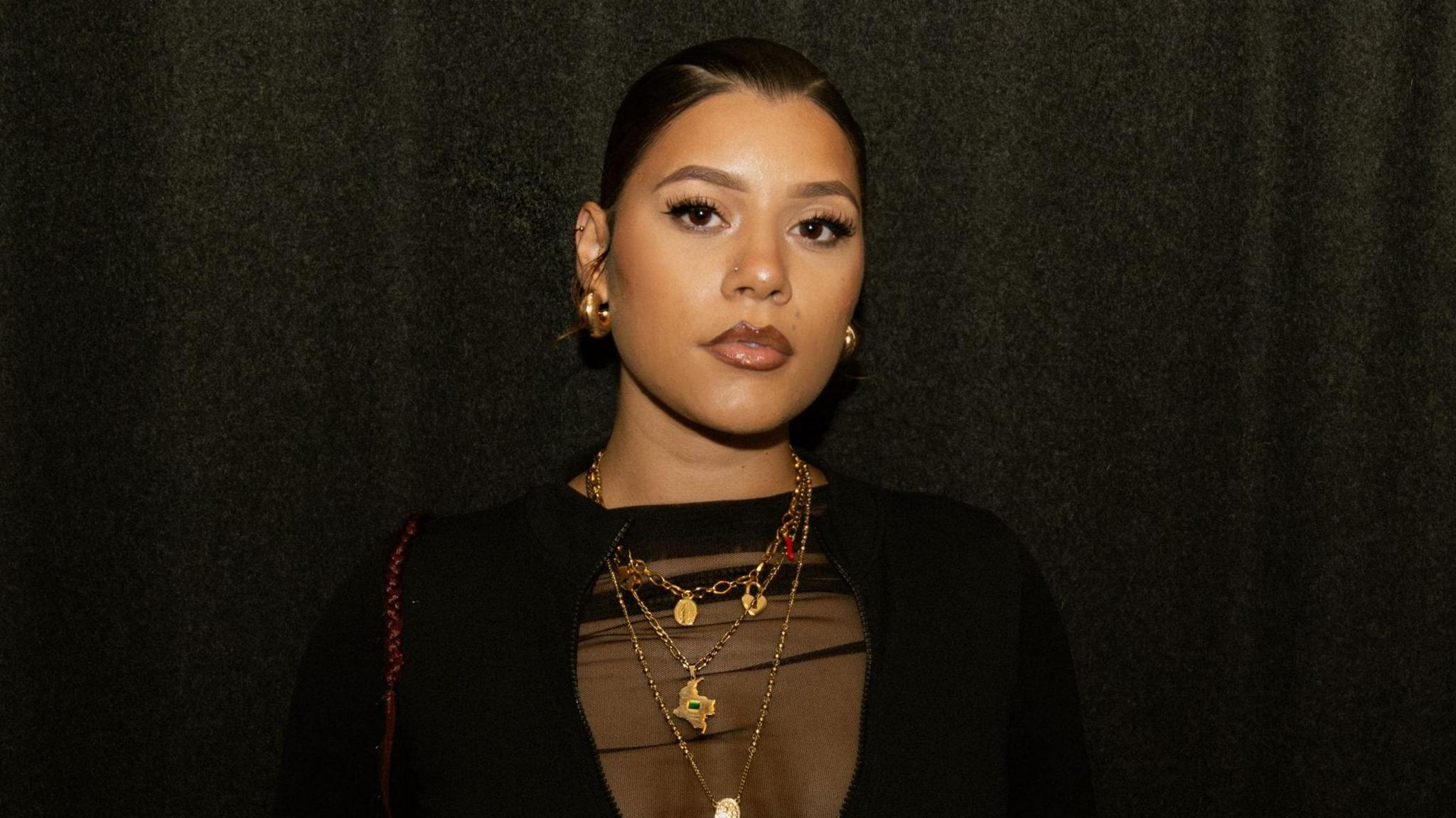 Milena Sanchez, pictured with her hair tied back in a slick bun. She looks at the camera with a serious expression, wearing a black sheer top and multiple gold necklaces. She's pictured inside against a dark backdrop. 