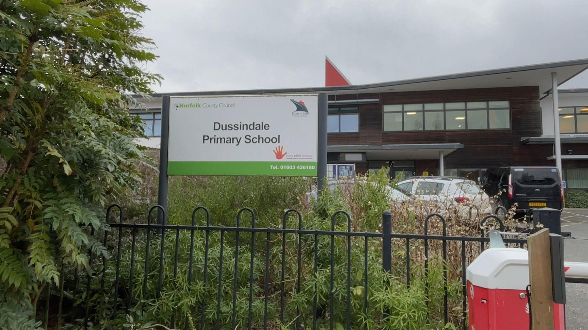 The exterior and entrance of Dussindale Primary School. The metal gates, some bushes and a school sign are visible. In the background is the school building, which has a wood-panelled effect.