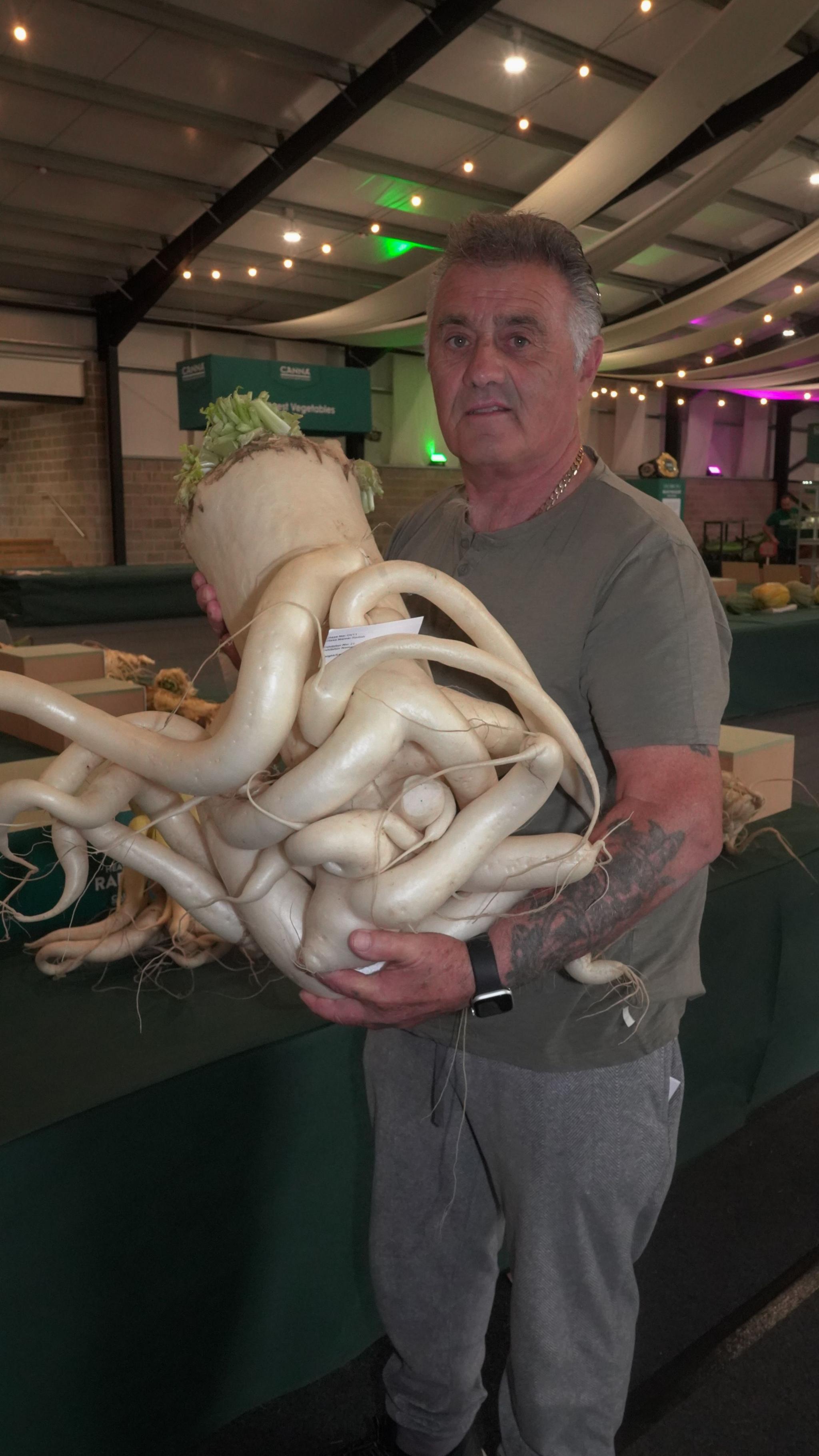 A man cradles a huge white root vegetable in his arms. The vegetable has limbs wrapping around itself. The man looks blankly into the camera. 