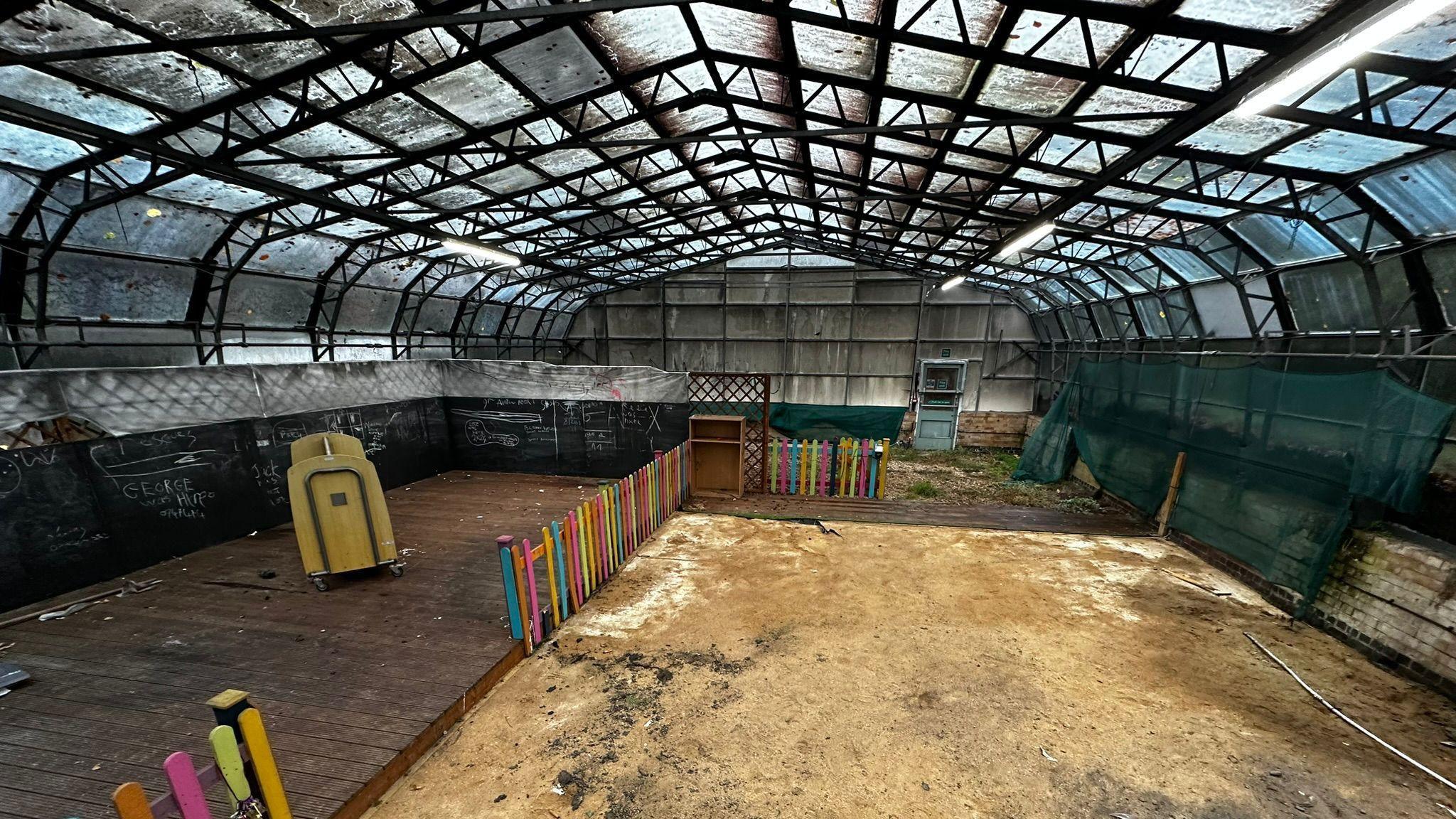 The interior of a large glass house. It has a glass-panelled roof and walls, upheld by a black complex steel structure. The glass panels look dirty. Most of the floor is covered with sand, but a raised area of wooden decking stands to the left, surrounded by a rainbow-coloured wooden fence.