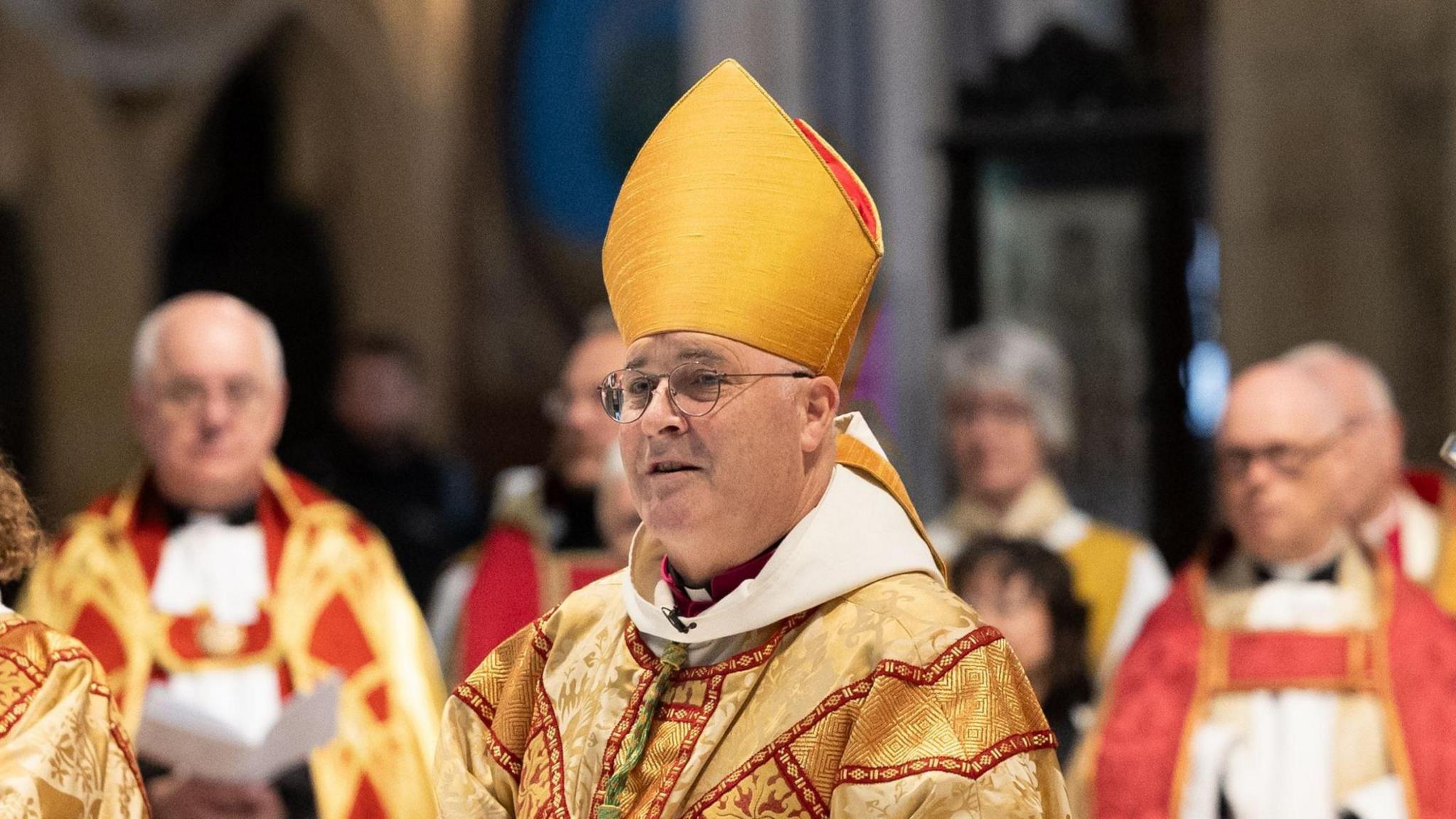 A man wearing round glasses stands in gold, red and white robes with a tall gold hat behind stand other members of the clergy. 