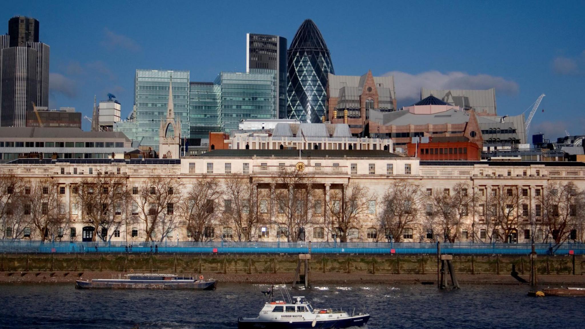 The front of the historic Custom House building viewed from the opposite side of the river. The building is behind some leafless trees, and is made of light-brown coloured stone. The River Thames is in the foreground, with boats running along it. The skyline of the City of London is visible in the background, with high-rise buildings including the Gherkin.