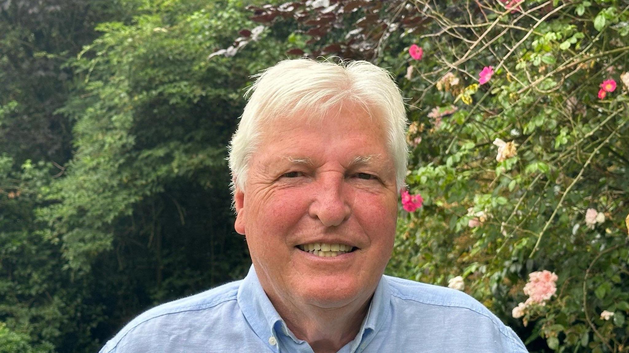 John Hayes in a blue shirt in smiling in front of roses while stood in garden.