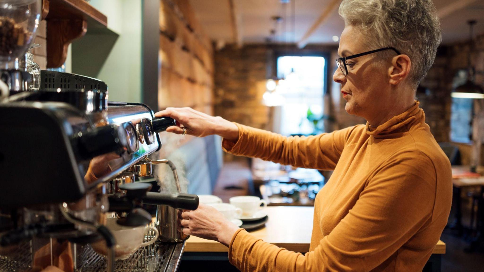 A woman with short grey hair and glasses wearing a yellow turtleneck, long-sleeved top steams milk in a professional coffee making machine