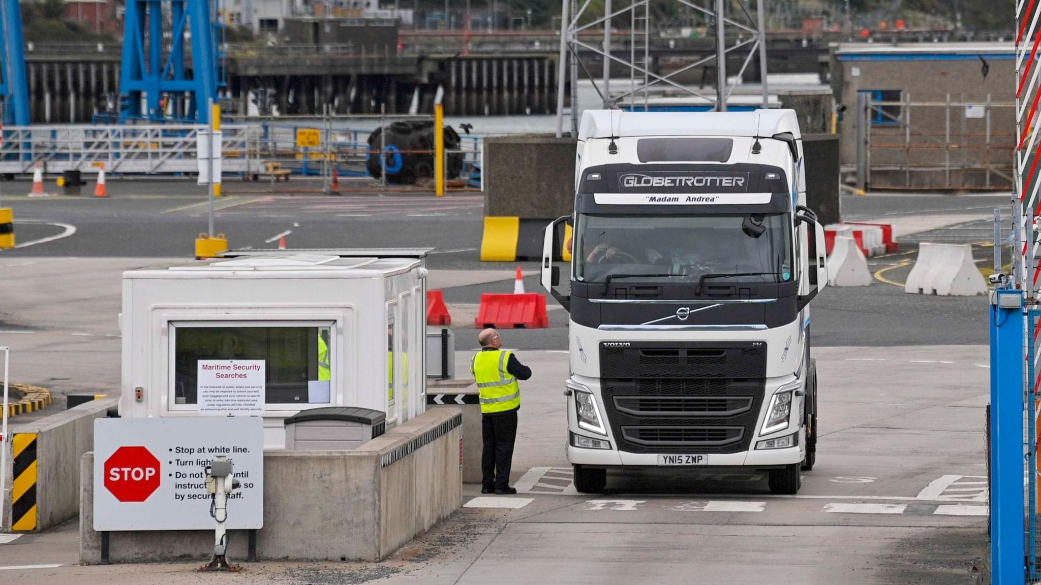 A wide shot of a white lorry leaving Larne Port. A driver is speaking to a port worker wearing a hi-vis jacket.