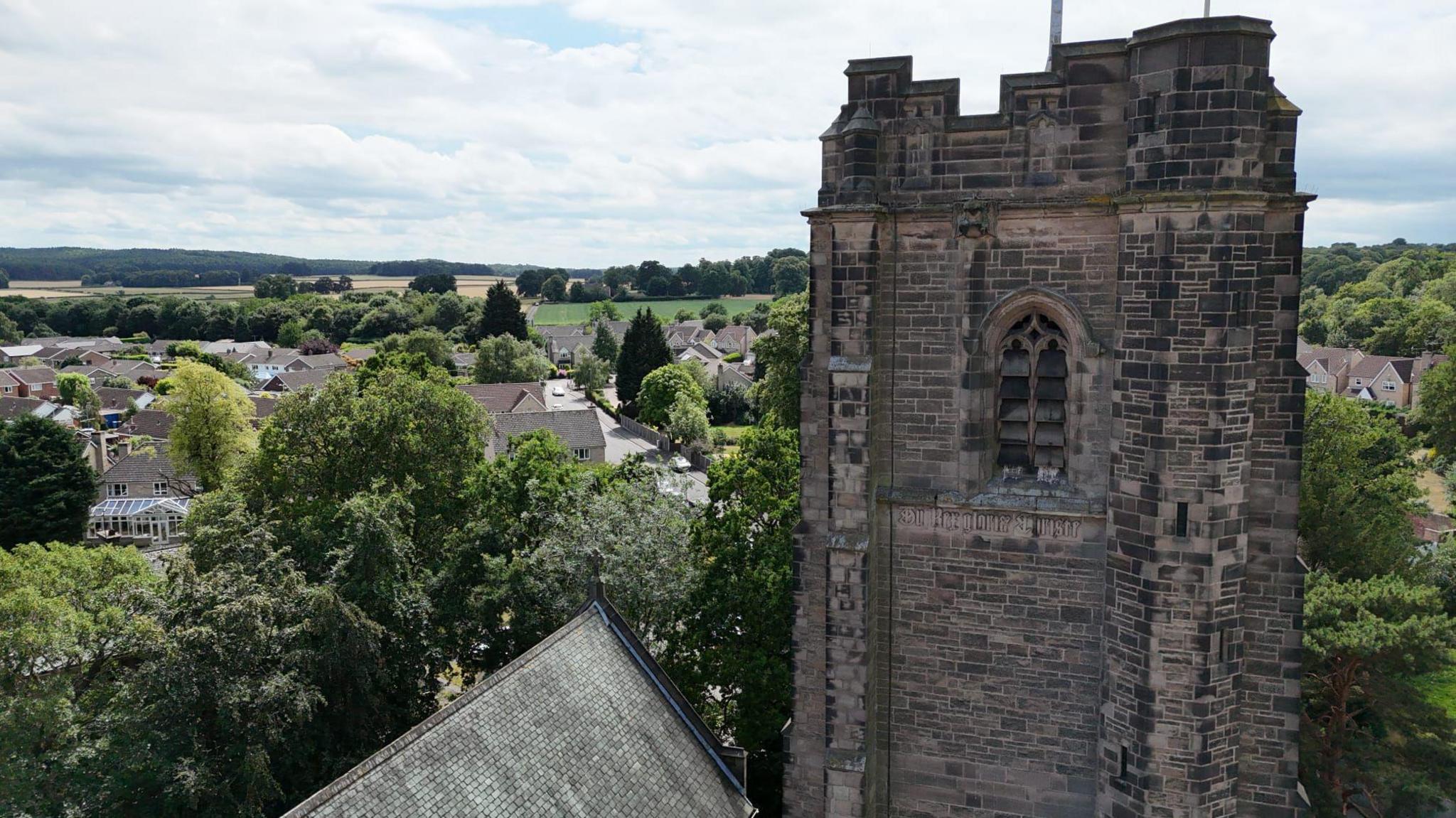 An aerial photograph of a church tower with house roofs and trees in the background
