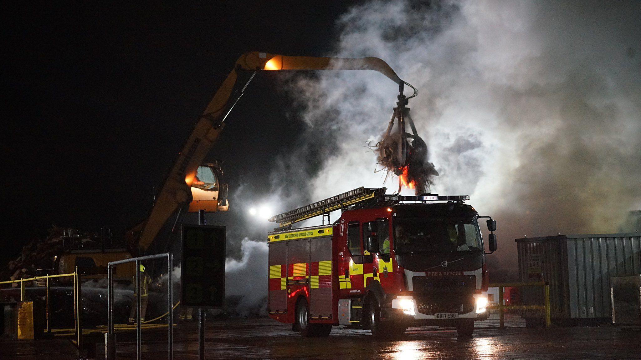 A fire engine in front of a fire. There is lots of smoke against a night sky. A crane is next to the fire engine lifting flaming materials into the air.