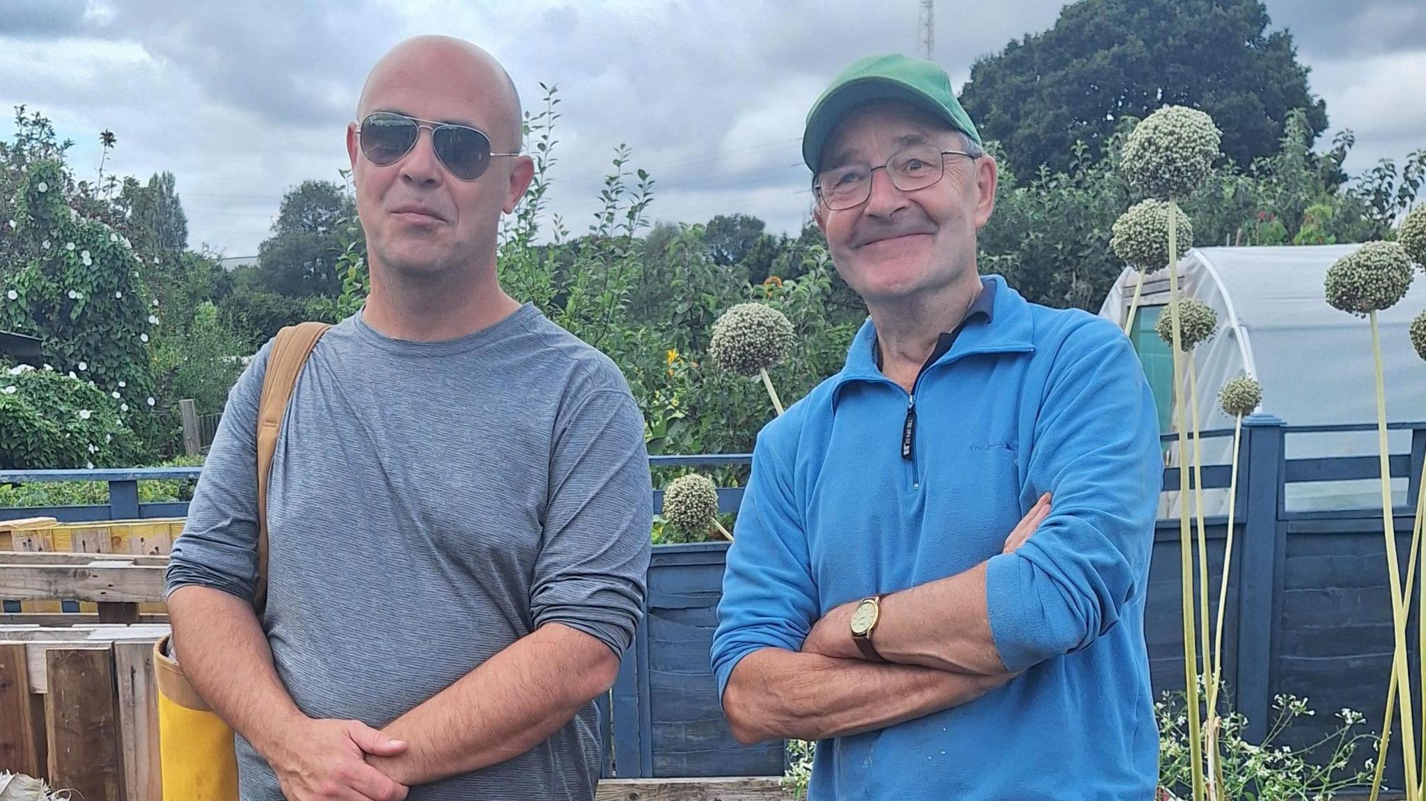 Two men stood side by side with the allotment behind them. The man on the left is wearing a grey top and sunglasses and folding his hands in front of him. The man on the right, who is a volunteer at the plot, is wearing a blue jumper, green cap and is folding his arms. 