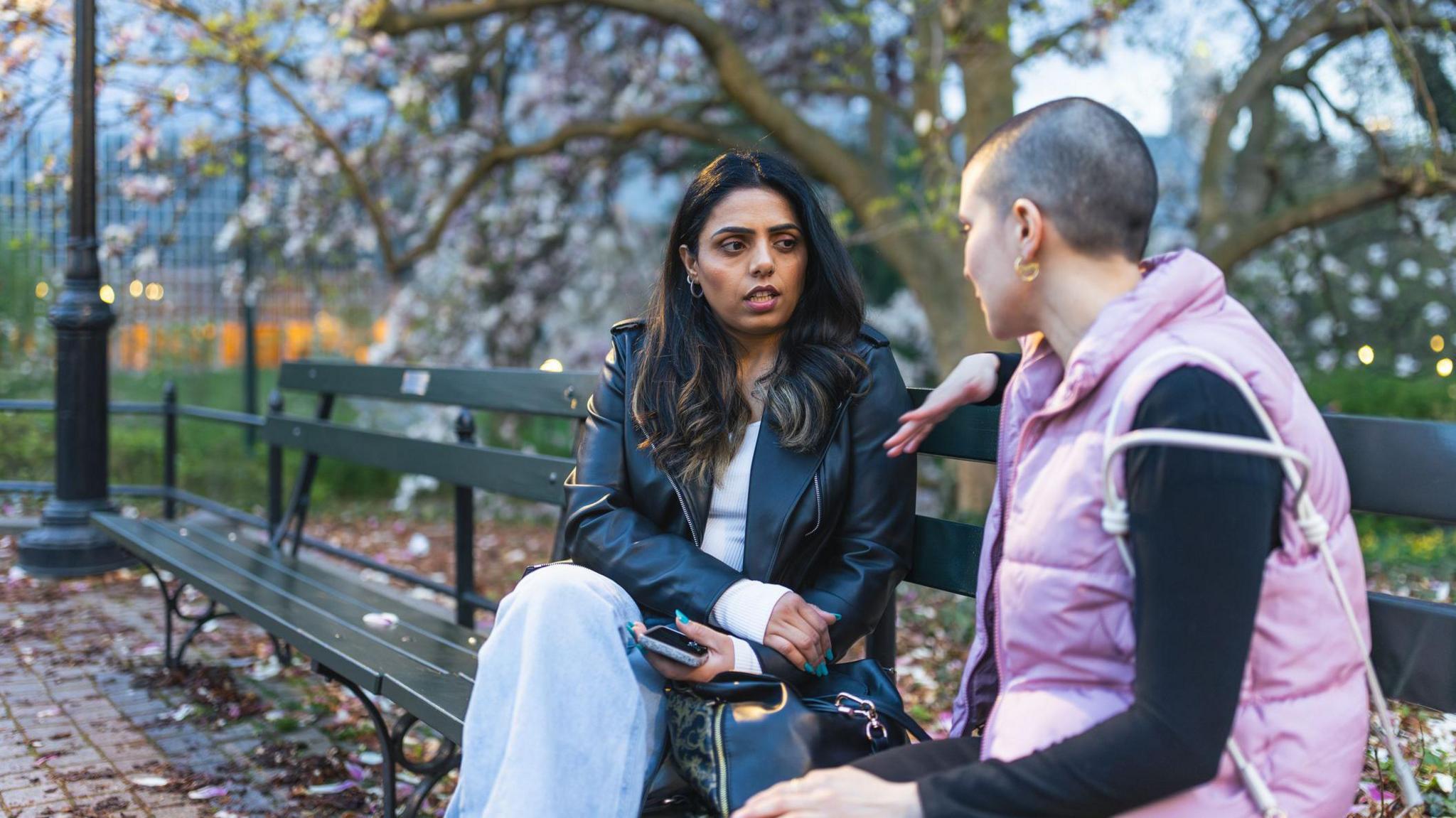 A woman with brown hair wearing a black leather jacket, white top and blue jeans and a woman with a shaved head wearing a black long-sleeved top and pink gilet sat on a brown bench. 