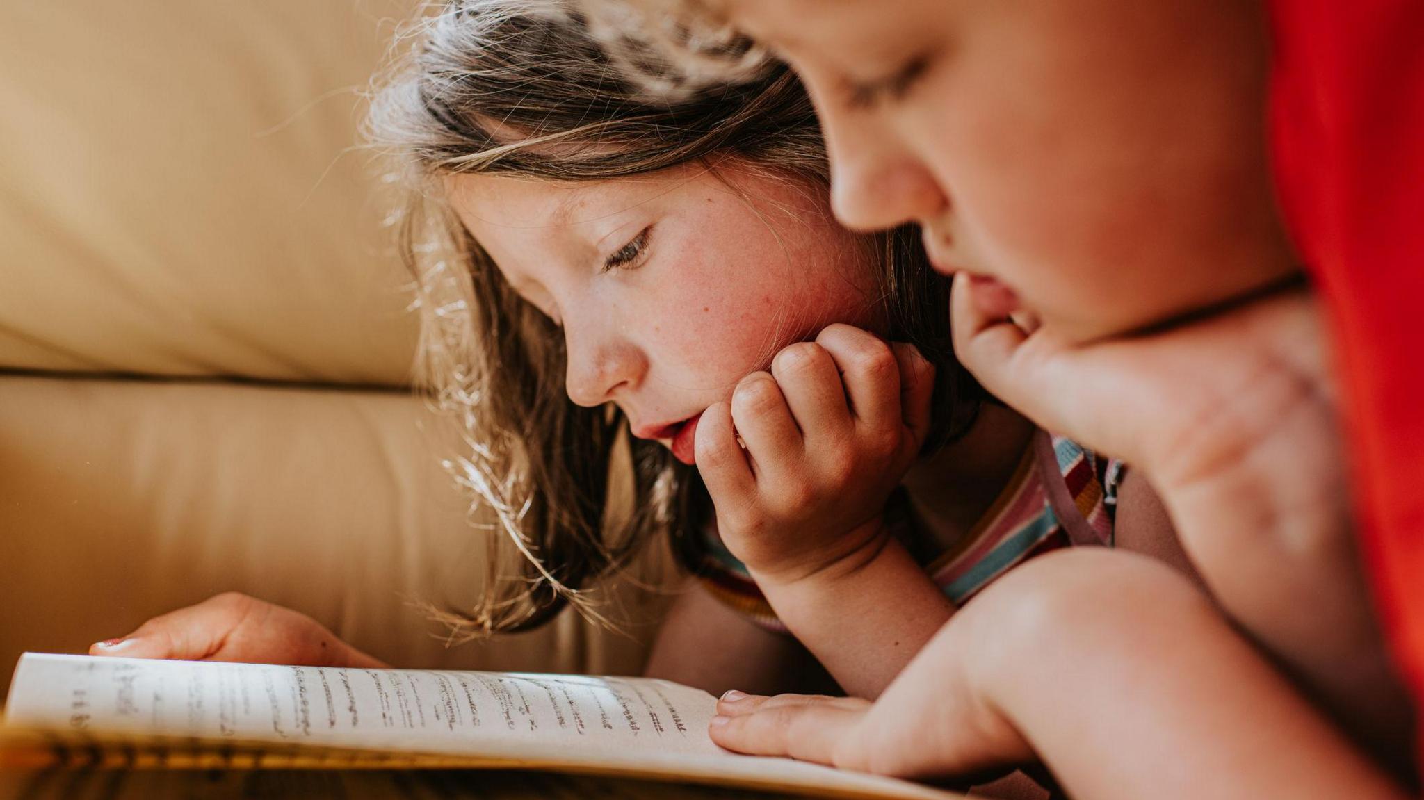 Two children are lying on their tummies, reading a book which is open in front of them.