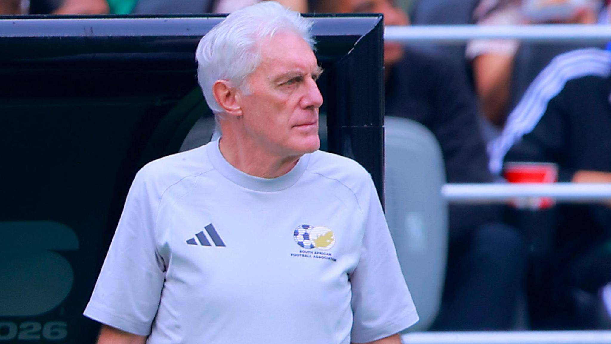 Hugo Broos, visible from waist up wearing a light grey South Africa t-shirt with federation crest and sportswear logo, looks to his left as he stands in front of a dugout at a World Cup match. A couple of members of the crowd can be seen out of focus behind him