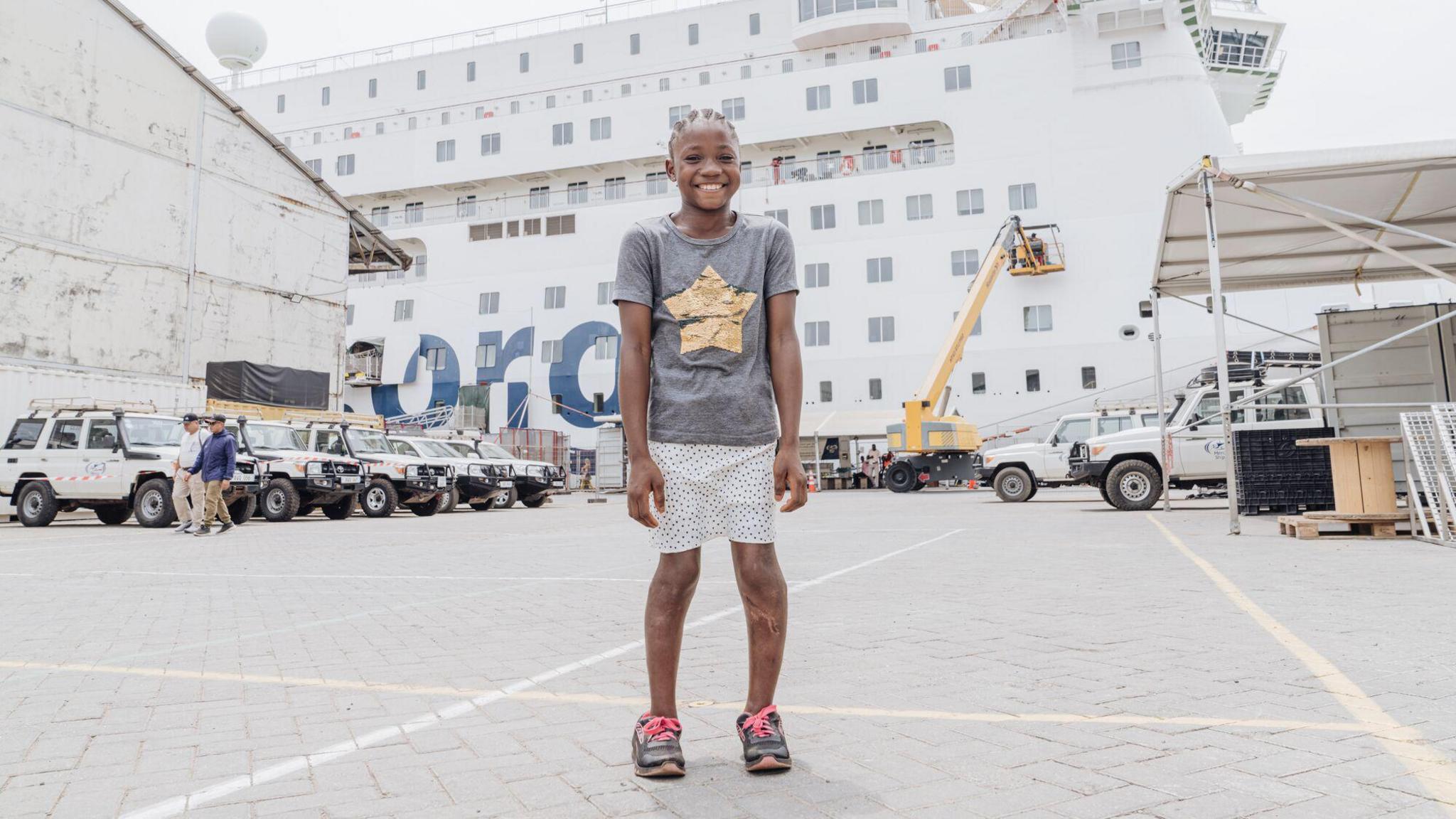 Fudia, after her surgery, she stands outside in a different section of the dockyard, with a white and blue Mercy Ship in the background, as well as a number of parked white jeeps. Fudia wears her hair in braids, with a slate grey short sleeved cotton t shirt and a gold foil star on the front, she has pale pink capri shorts and wears black platform sliders. She is smiling, with her legs straighter and now wears black trainers with neon pink laces.