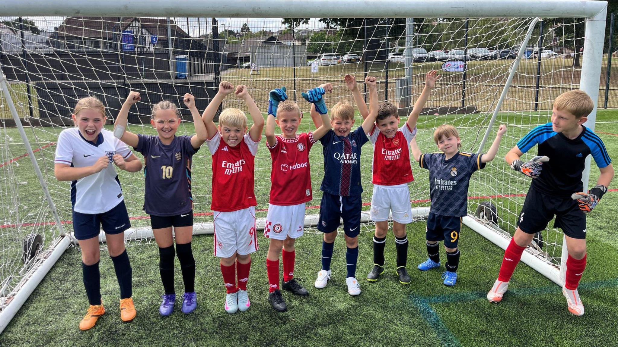 A group of young children in football kits, six boys and two girls, wave and cheer as they stand inside a goal at a Bristol City Football Club summer event.