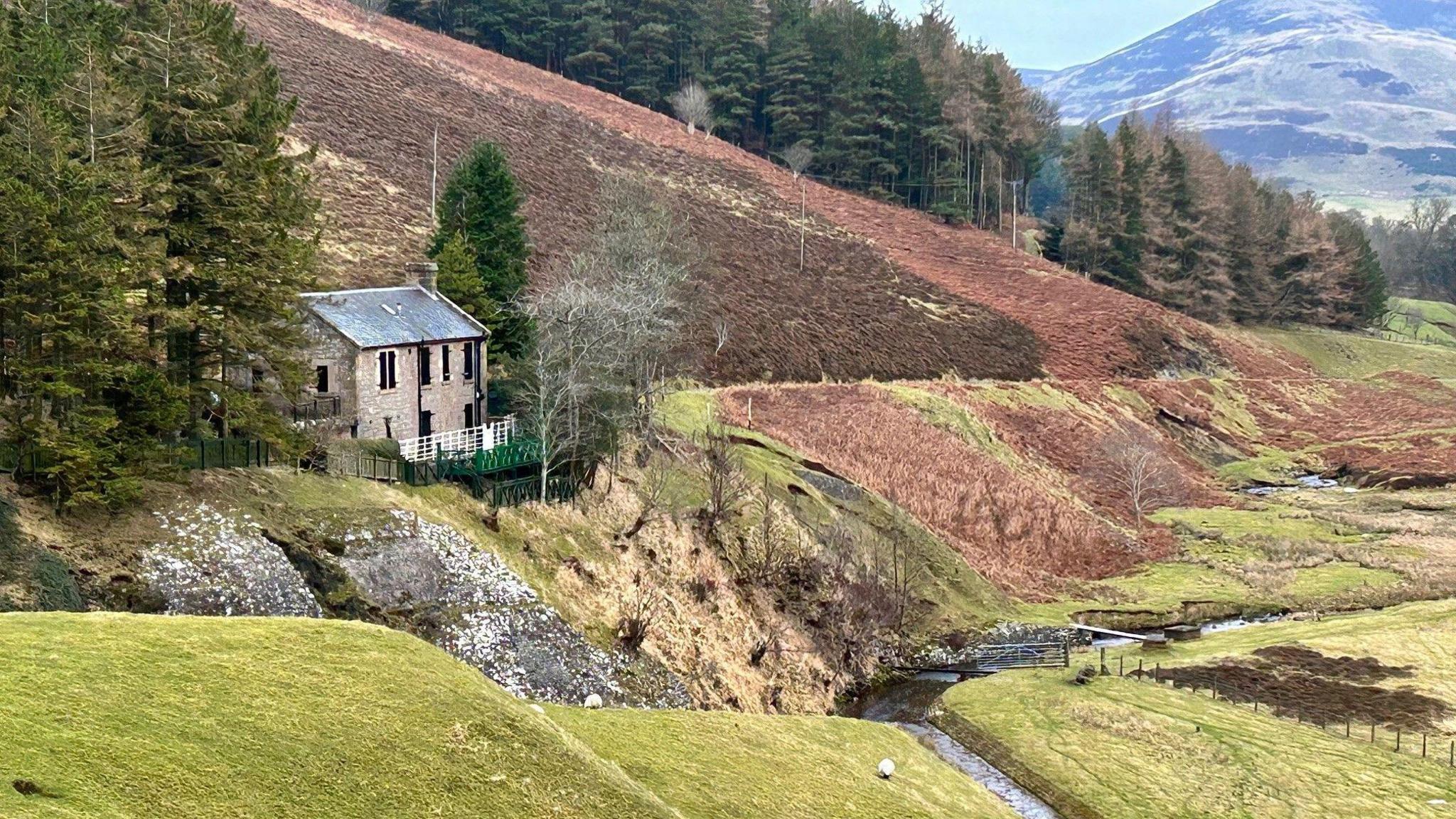 An old two-storey building is on the left of the picture. It is midway down a picturesque hillside. There are conifer trees in the background and burn in the foreground. 