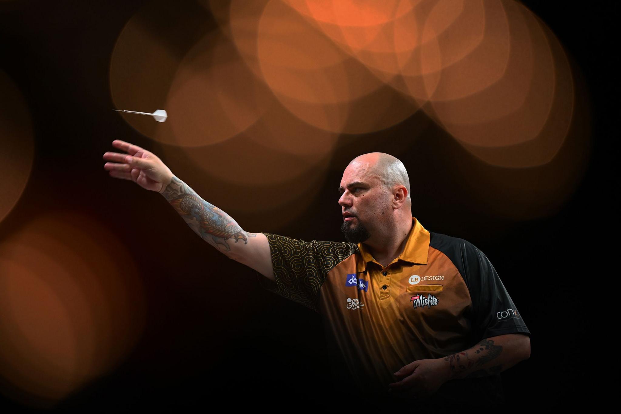 Person throwing a dart during a game, wearing a black and orange patterned shirt with visible tattoos on the arm, against a dark background with blurred circular lights.