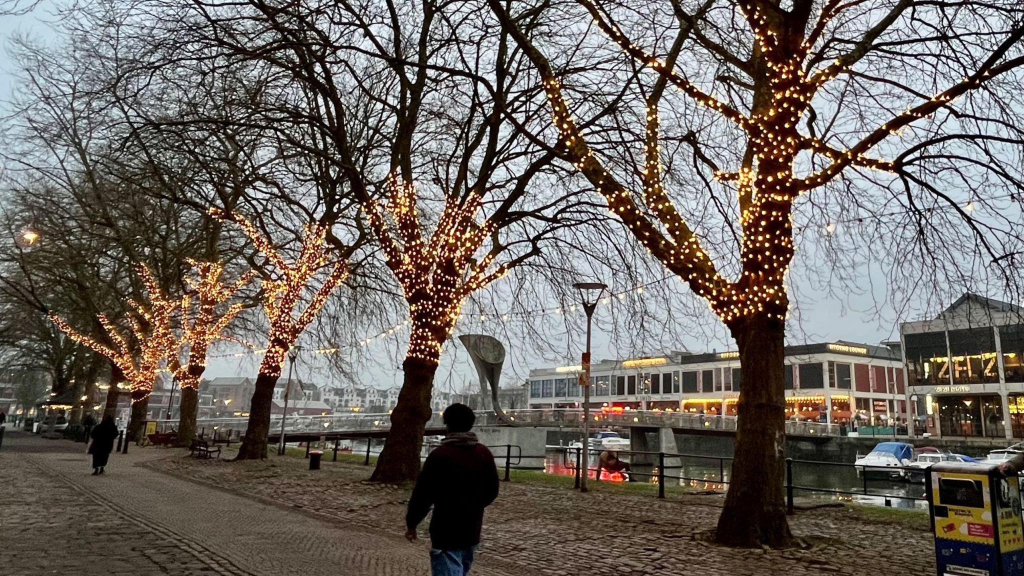 A man in dark clothing walks along one side of the Bristol harbourside opposite a row of brighly-lit restaurants and bars. He is walking past five large trees which have hundreds of small lights attached to their trunks and lower branches