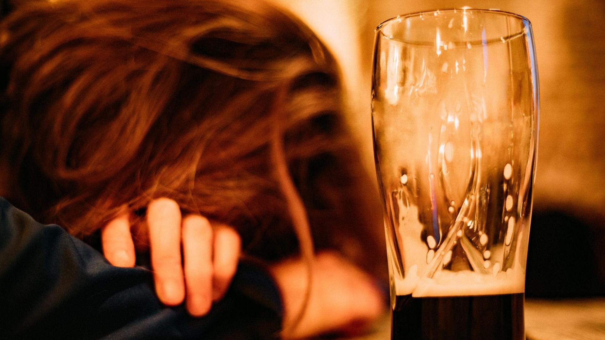 A woman with ginger hair sat with her head in her arms appearing asleep and drunk on a bar. An almost empty pint of dark beer is next to her. Stock picture.