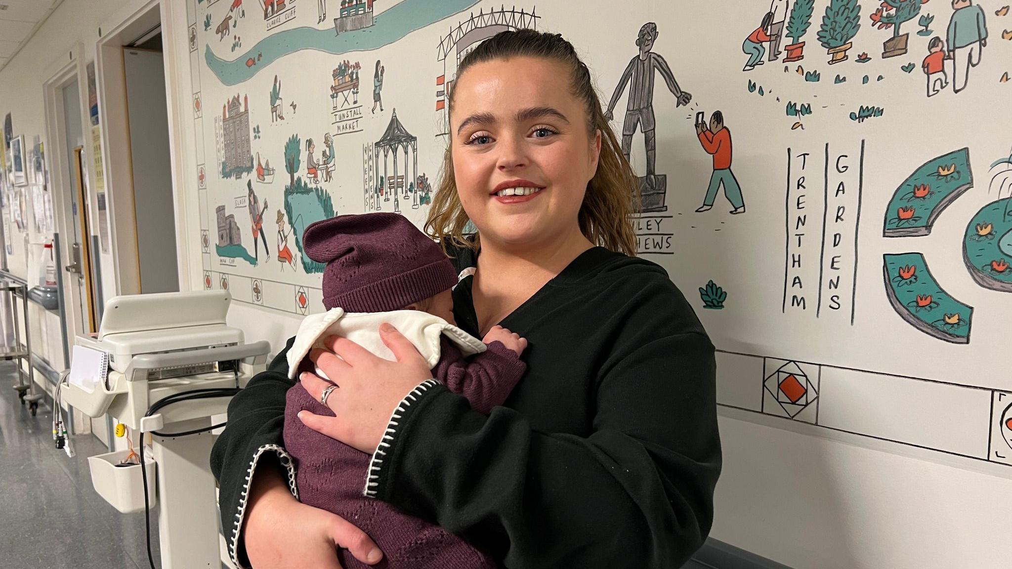 Natasha, wearing a black jumper, is holding her baby. The baby is wearing a maroon hat and baby-gro. They are in a hospital corridor.