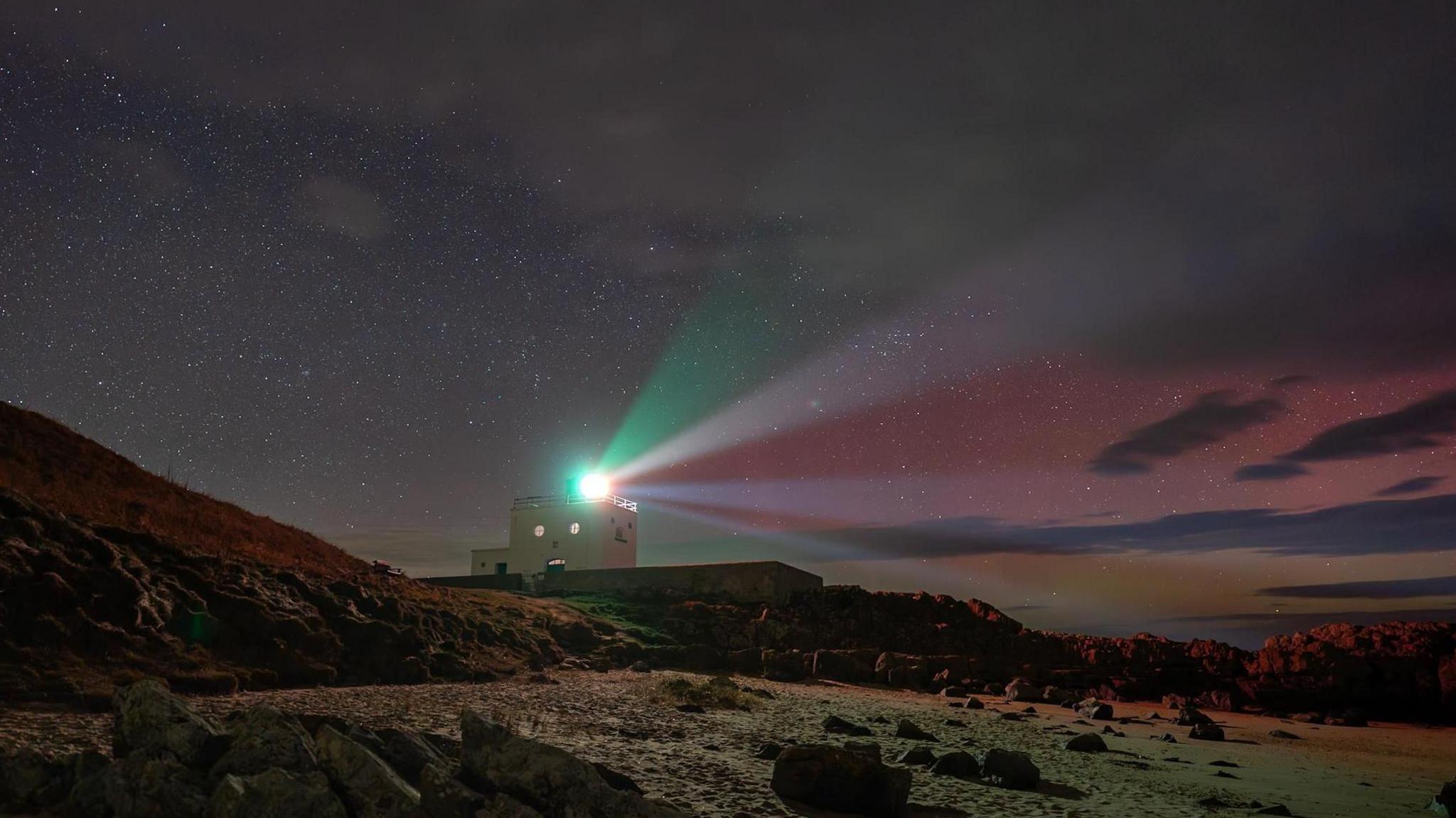 Bamburgh lighthouse with beams of light shining out in the night sky with the aurora showing pink and orange colours in the sky.