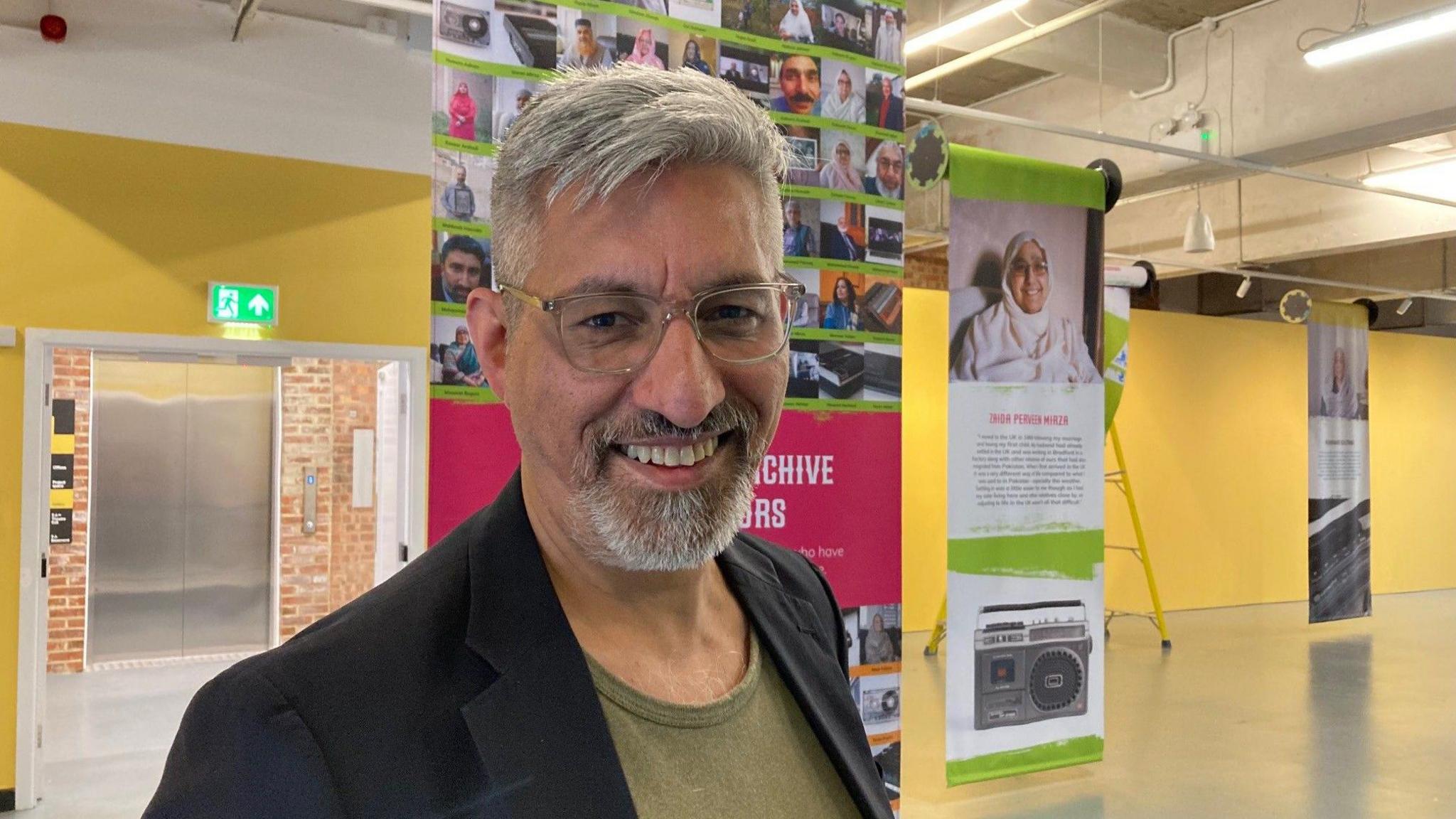 A silver-haired man man wearing glasses who is smiling while standing in front of a couple of posters hanging from a ceiling.