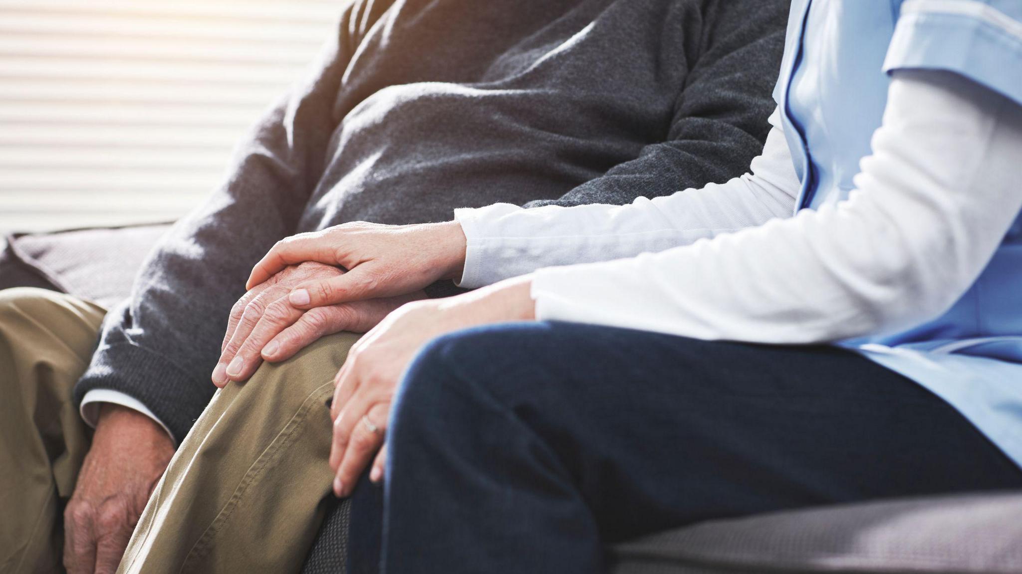 Close up of a care workers hand on top of an elderly man's hand. The care worker is wearing a light blue tunic and the man is wearing brown trousers and a grey jumper. Their faces are not shown.