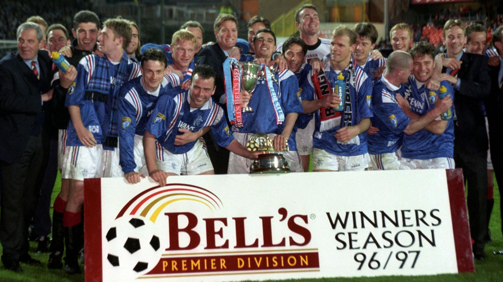 Rangers football team wearing blue shirts and white shorts standing in front of a banner on the pitch that reads BELL'S PREMIER DIVISION WINNERS SEASON 96/97. The team are celebrating and holding two trophies.
