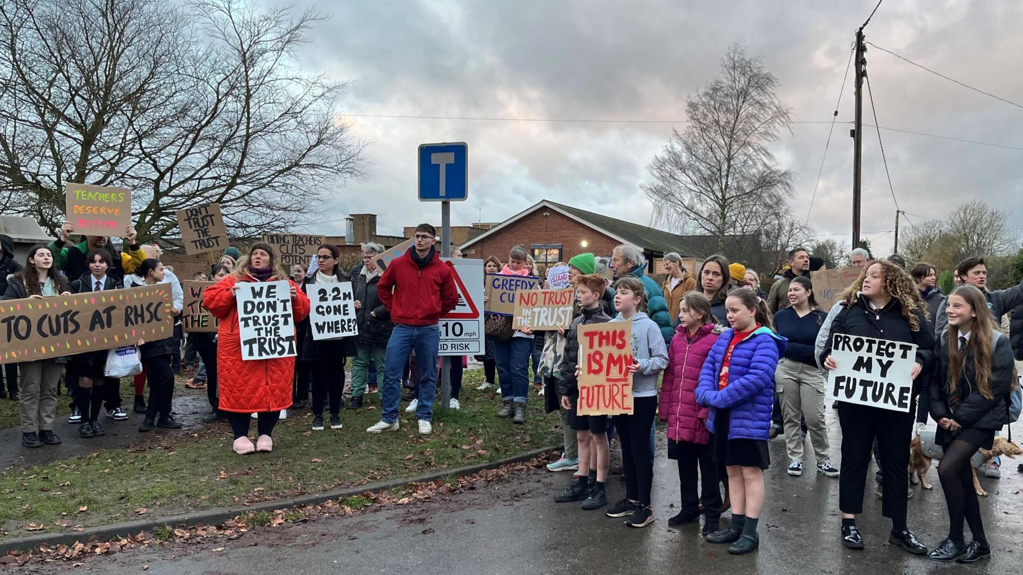 Some 50 or so adults and children - some holding placards - stand outside a school. The placards are hand-made out of cardboard and carry messages opposing school staff cuts.