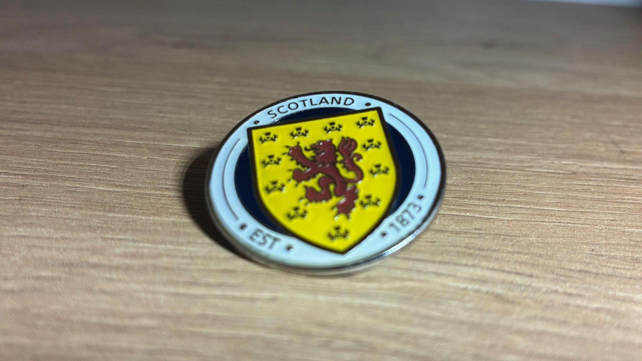 A circular metal badge with the Scotland crest showing a red lion rampant on a yellow background upon a navy circle with a white border. Text around the outside reads "Scotland Est 1873"
