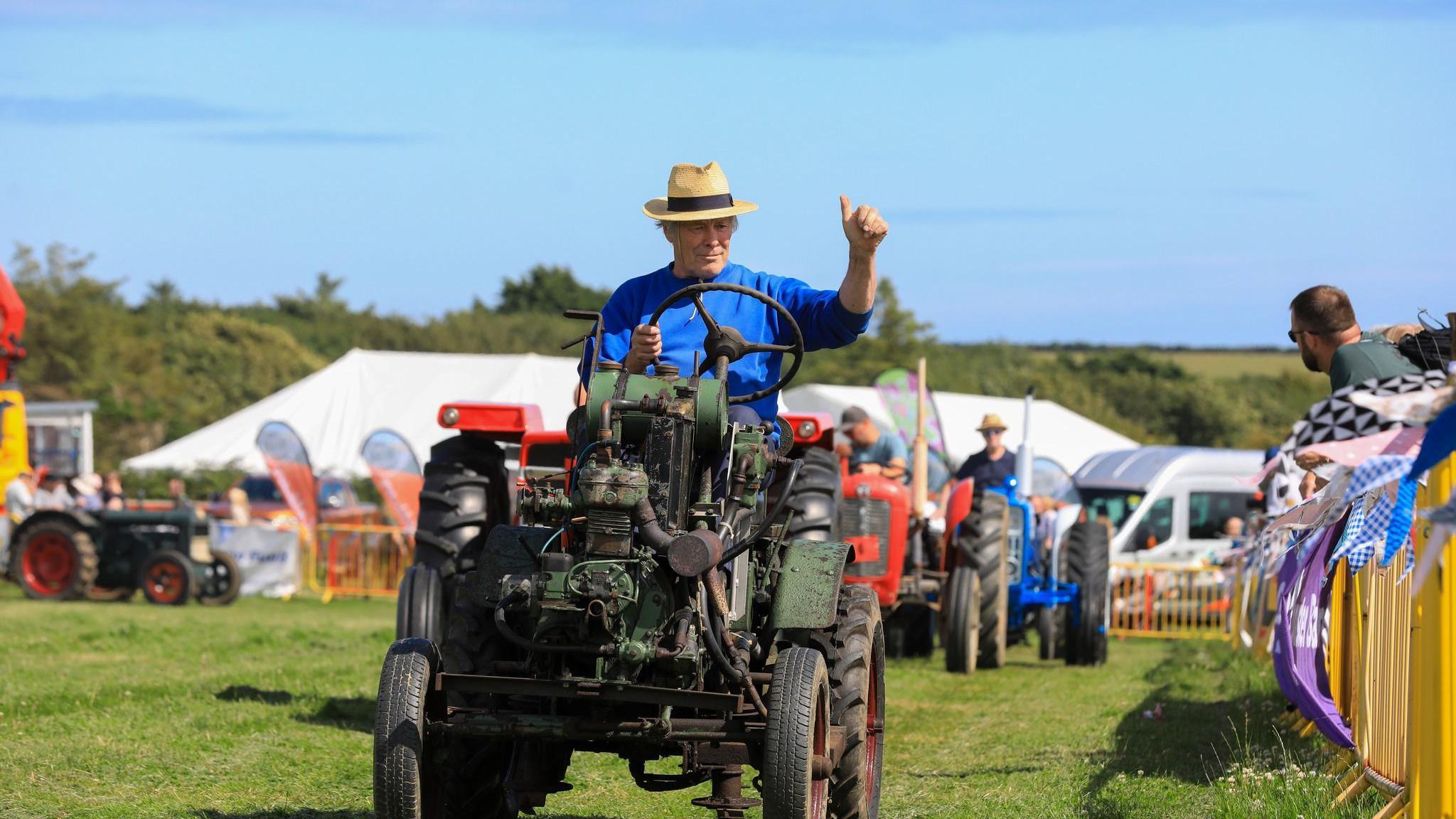 A man driving an old fashioned tractor wearing a trilby hat and giving the watching a thumbs up. He is leading a parade of other tractors in the grassed show ring.