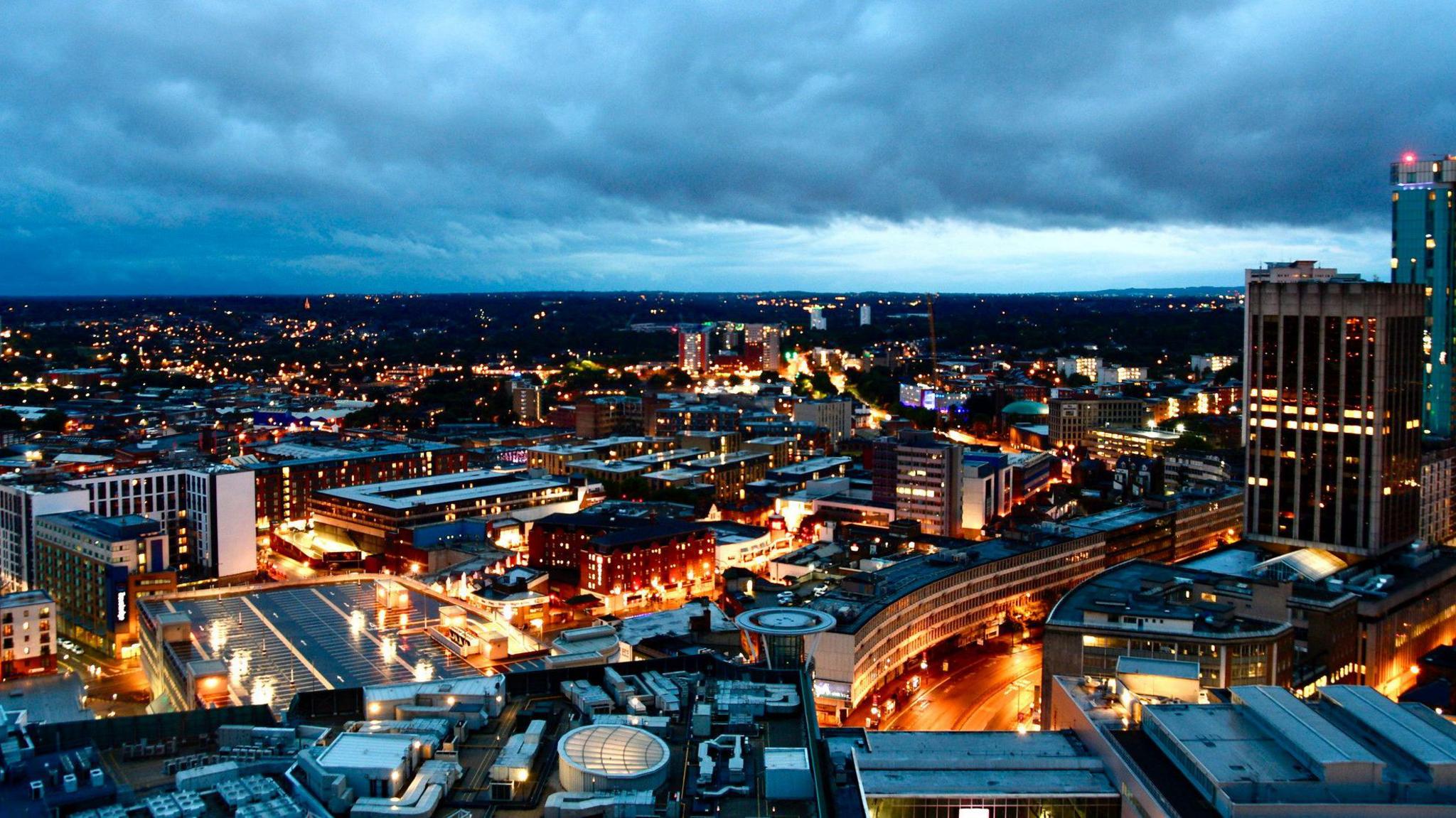 A view of Birmingham at night time. Buildings are lit up with orange/yellow lights. There is a high-rise building on the right which has a red light on the top of it. The building stretch into the background where it is darker