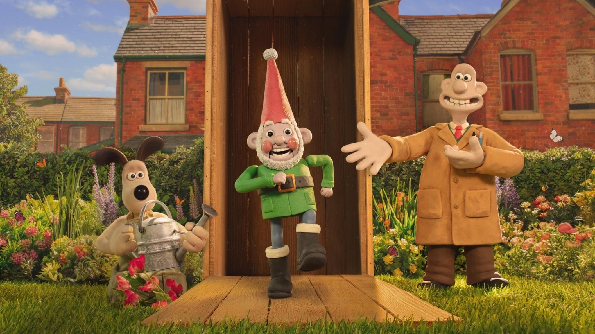 A production still from the film showing Wallace, Gromit and Norbot the gnome. The characters are in a garden setting. The gnome is walking on the spot in front of a shed, Gromit is holding a watering can