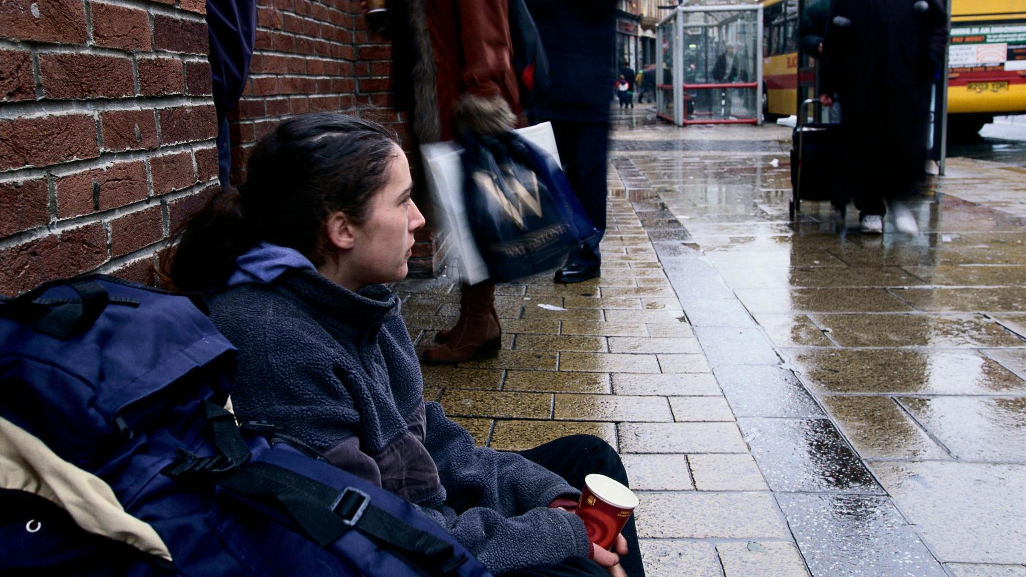 A woman sitting on the pavement with her back against a red brick wall and a rucksack beside her. She is holding a disposable coffee cup. In the background there are people walking past and a bus stop with a bus at it.