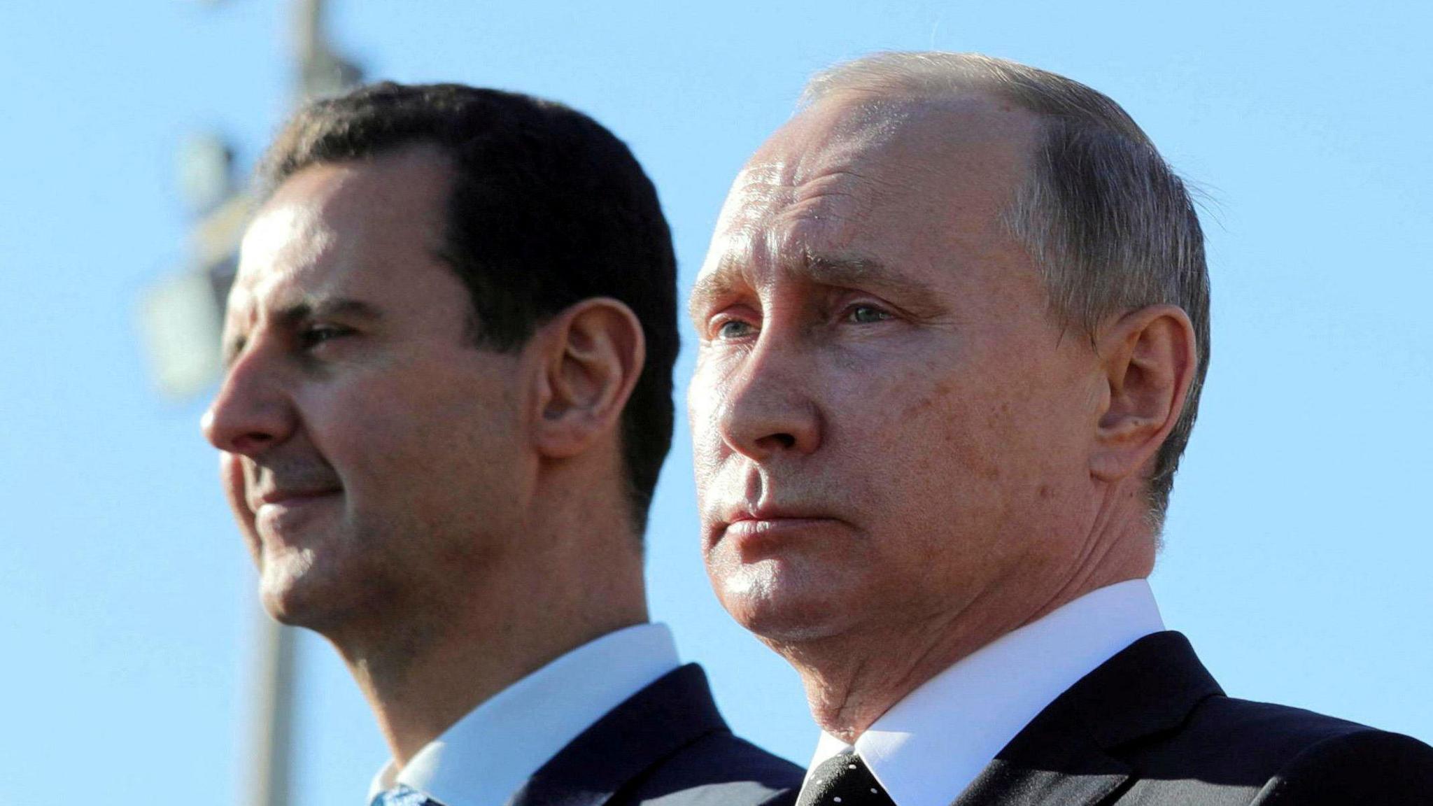 Putin and Assad stand next to each other. Both are wearing suits, with the sky acting as background