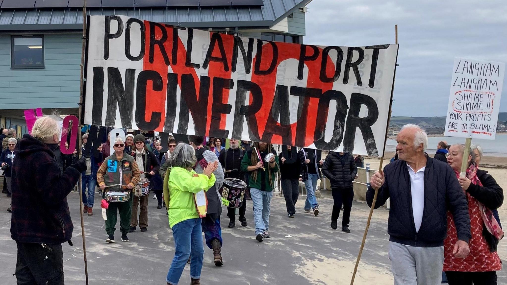 Protesters carrying signs and drums walk beside a beach. A prominent banner reads "No Portland Port incinerator".