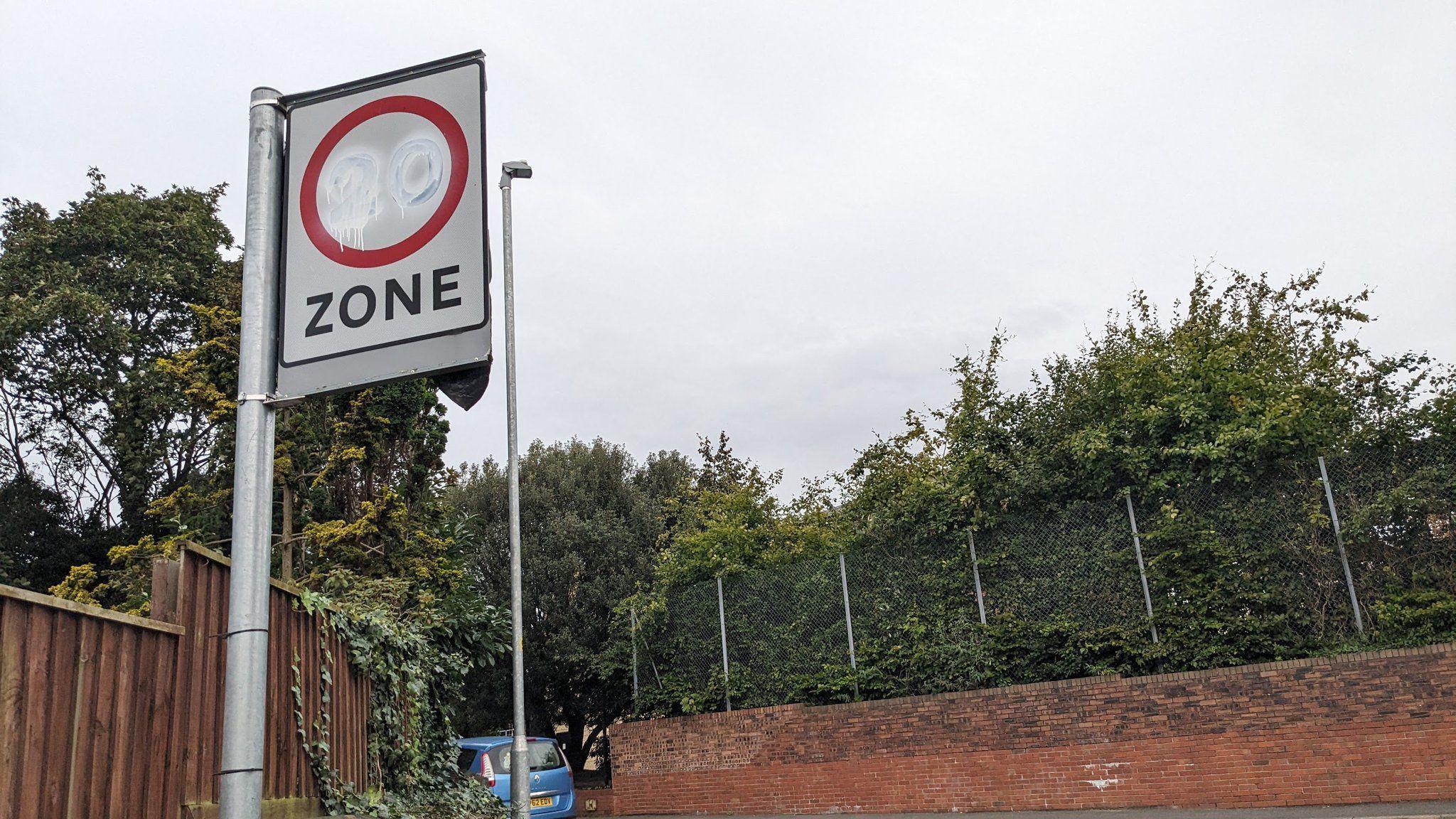 A 20mph speed limit sign on a suburban road is daubed with white paint, rendering the number 20 harder to see. There is a wall, fencing and trees in the background