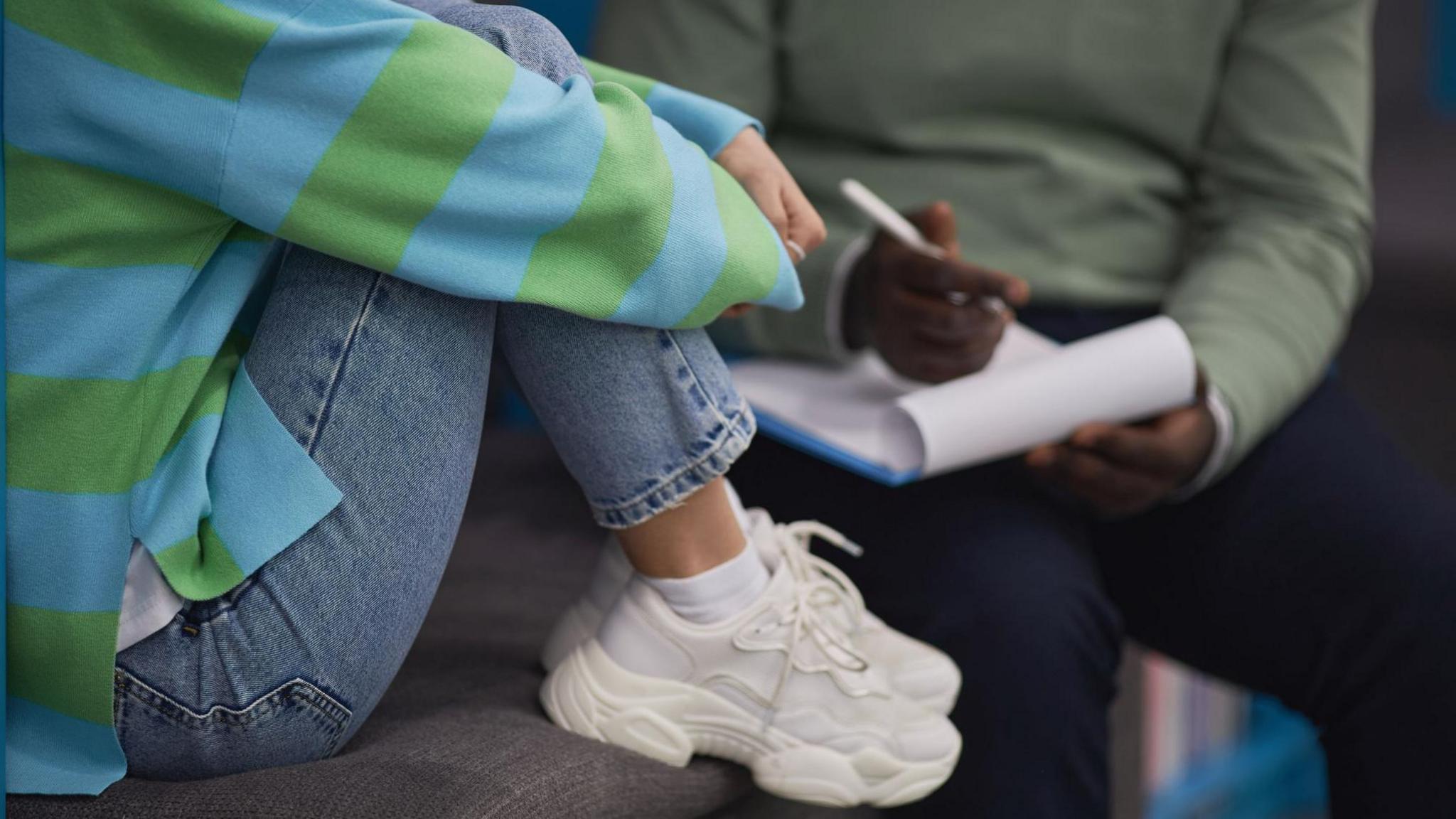 A shot of a teen in jeans, a green and blue striped jumper and white trainers sits with their feet up on a seat. A therapist in the background is holding a pen and note pad - he is wearing a green-grey jumper.