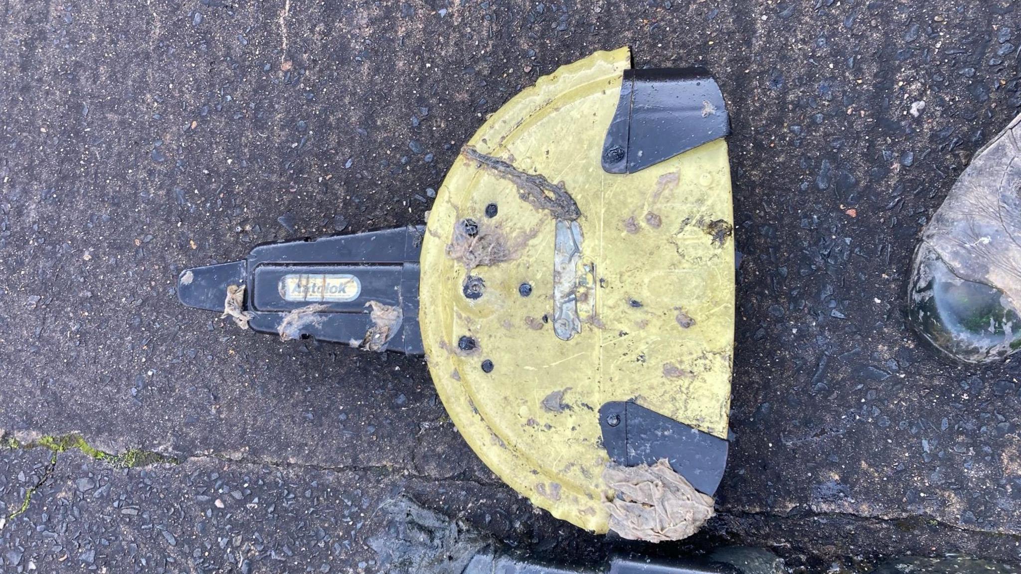 A steering wheel lock, laid on the ground. It is yellow and black. It is dirty, having been recovered from a sewer.