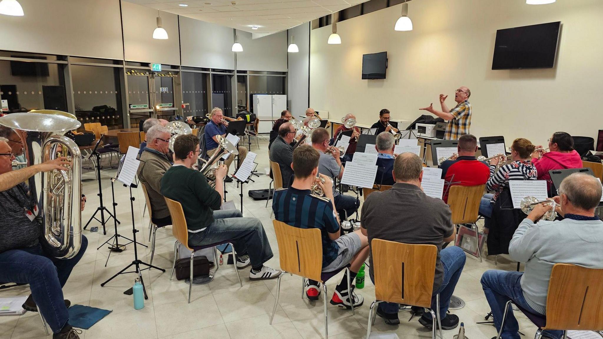 A brass band in rehearsal. About 20 people sit in a semi-circle in brown wooden chairs facing a conductor. They are playing instruments with music stands in front of them.