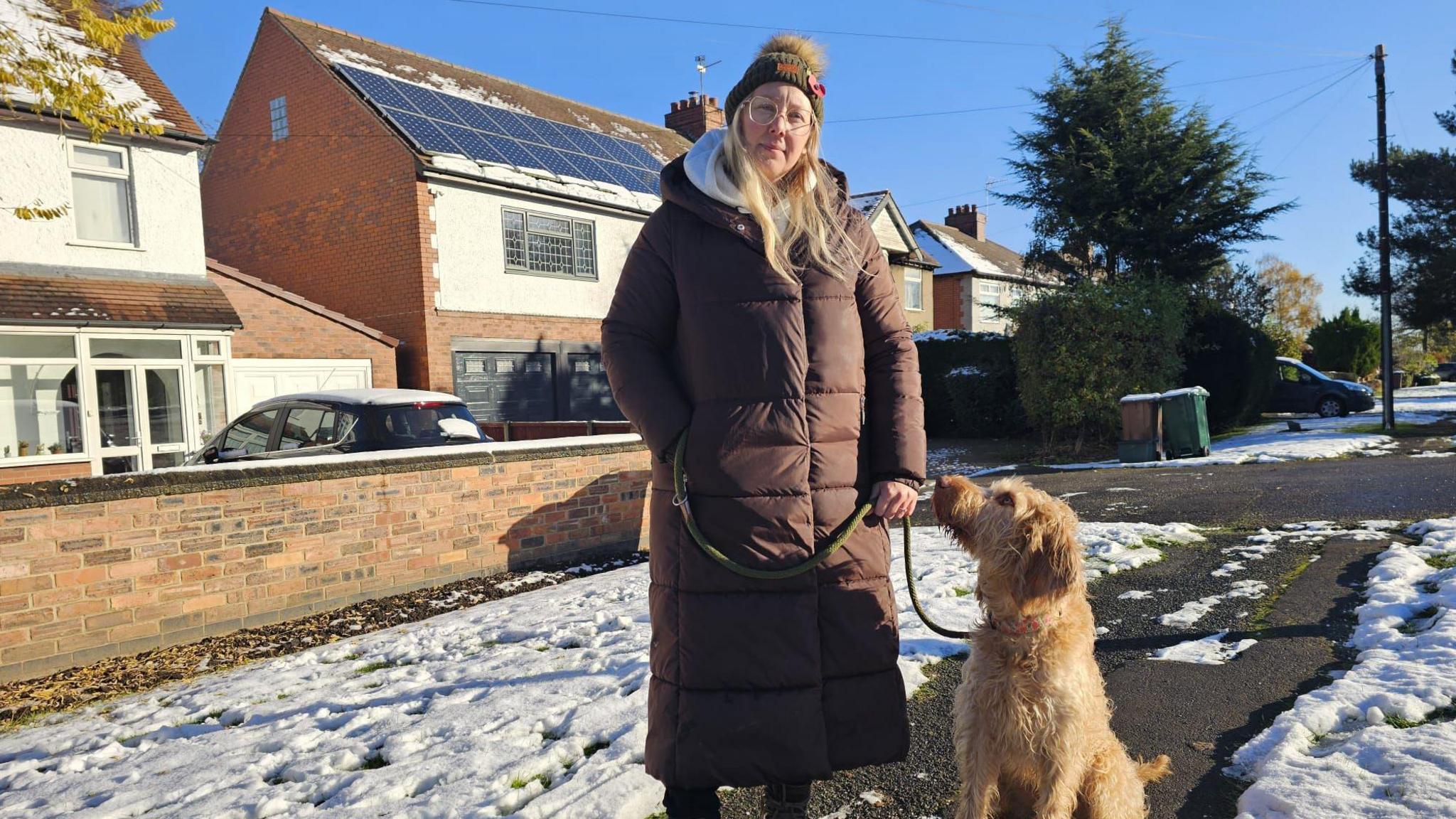 Katie Castle stands in front of her house with her dog, Peanut. She is wearing a long brown coat and a woolly hat with a bobble on. 